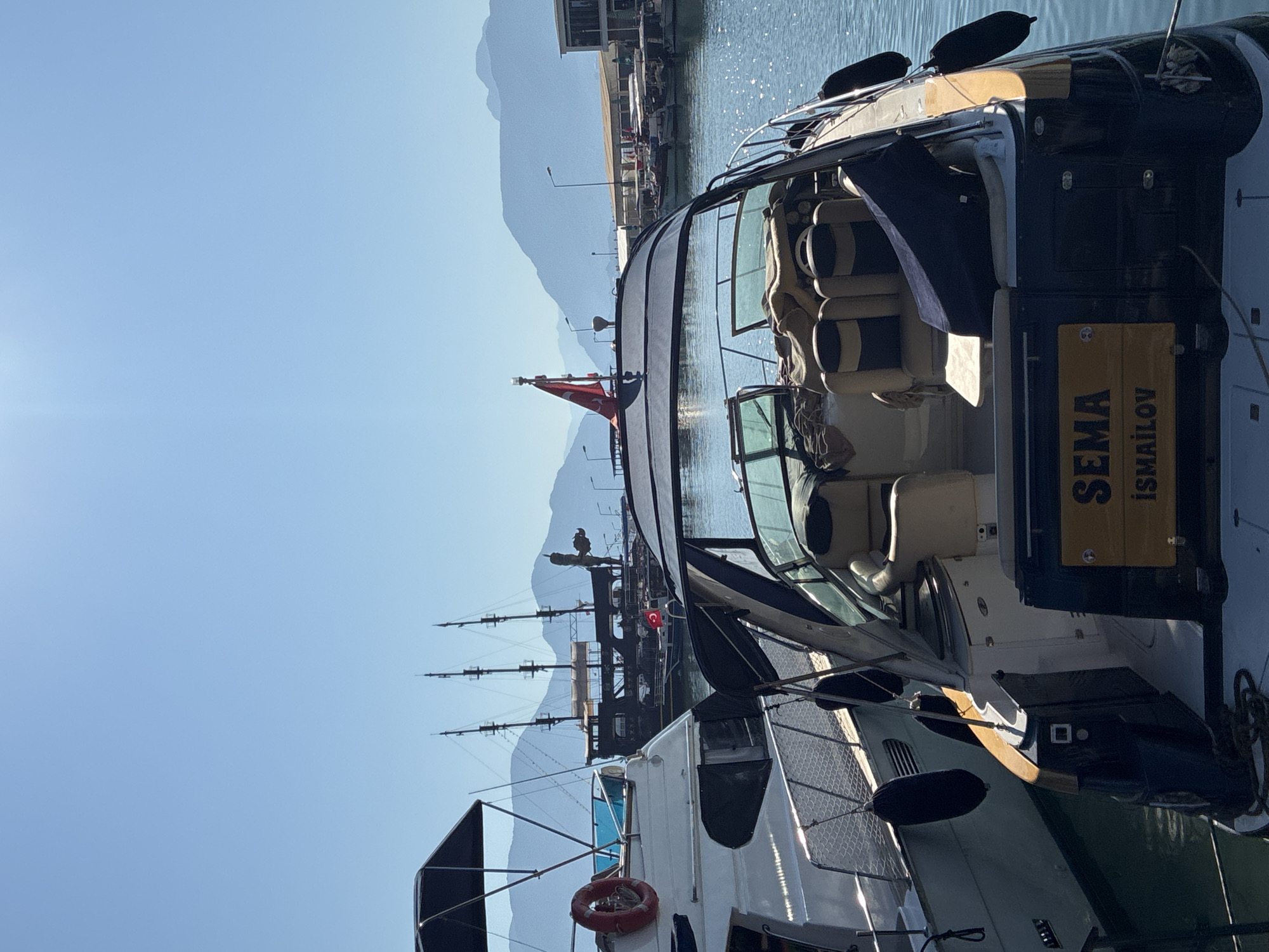 In Alanya, Turkey, a sun-drenched harbor scene unfolds under a clear blue sky. The foreground is dominated by the stern of a dark-hulled motor yacht, its nameplate prominently displaying "SEMA İSMAİLOV" in yellow lettering. The yacht features comfortable seating, a canvassed roof, and several black fenders attached to its sides. To the left, a white yacht with a dark blue trim is docked, also equipped with black fenders and a red life preserver. In the background, a striking, multi-masted ship with a dark hull and a figurehead is moored. Beyond the ships, the city of Alanya is visible, with buildings lining the waterfront, and beyond that, a range of hazy mountains under the bright sun. The water is calm and reflects the sunlight. No people are visible in the image.