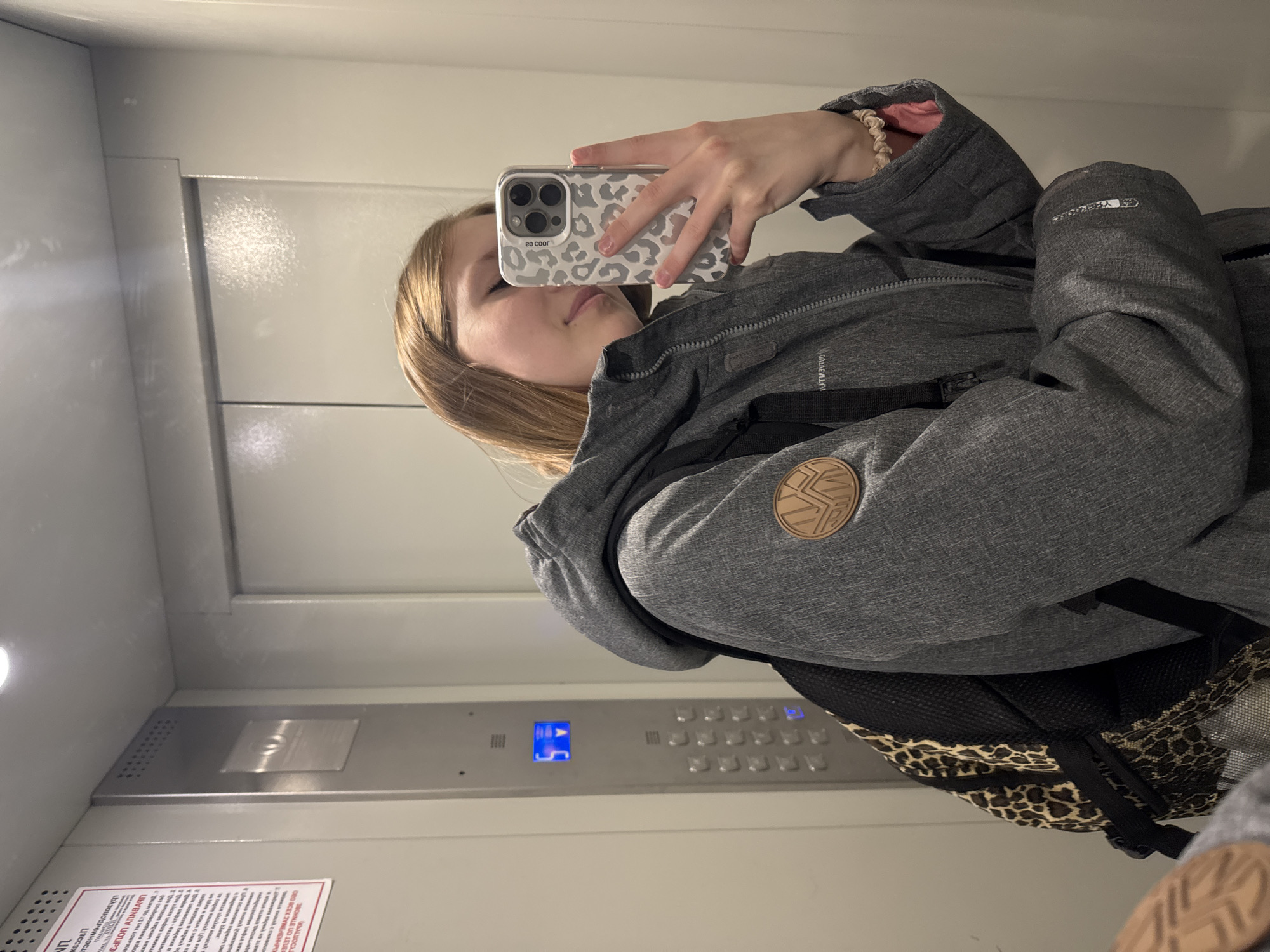 A young woman is taking a selfie in an elevator. She is wearing a gray jacket with a hood and carrying a backpack with a leopard print design. The elevator has a control panel with blue illuminated buttons. A sign with Russian text is visible on the bottom left of the elevator wall. The control panel indicates the elevator is on the 5th floor. The sign's text reads in part 'KALUOCA', 'СЕХ ДУЖЕЛЕННРО', 'ΠΝΒΑ', 'Εт од метад'. Based on the text of the sign, it is likely that this photo was taken in the city of Chaikovsky, Russia.