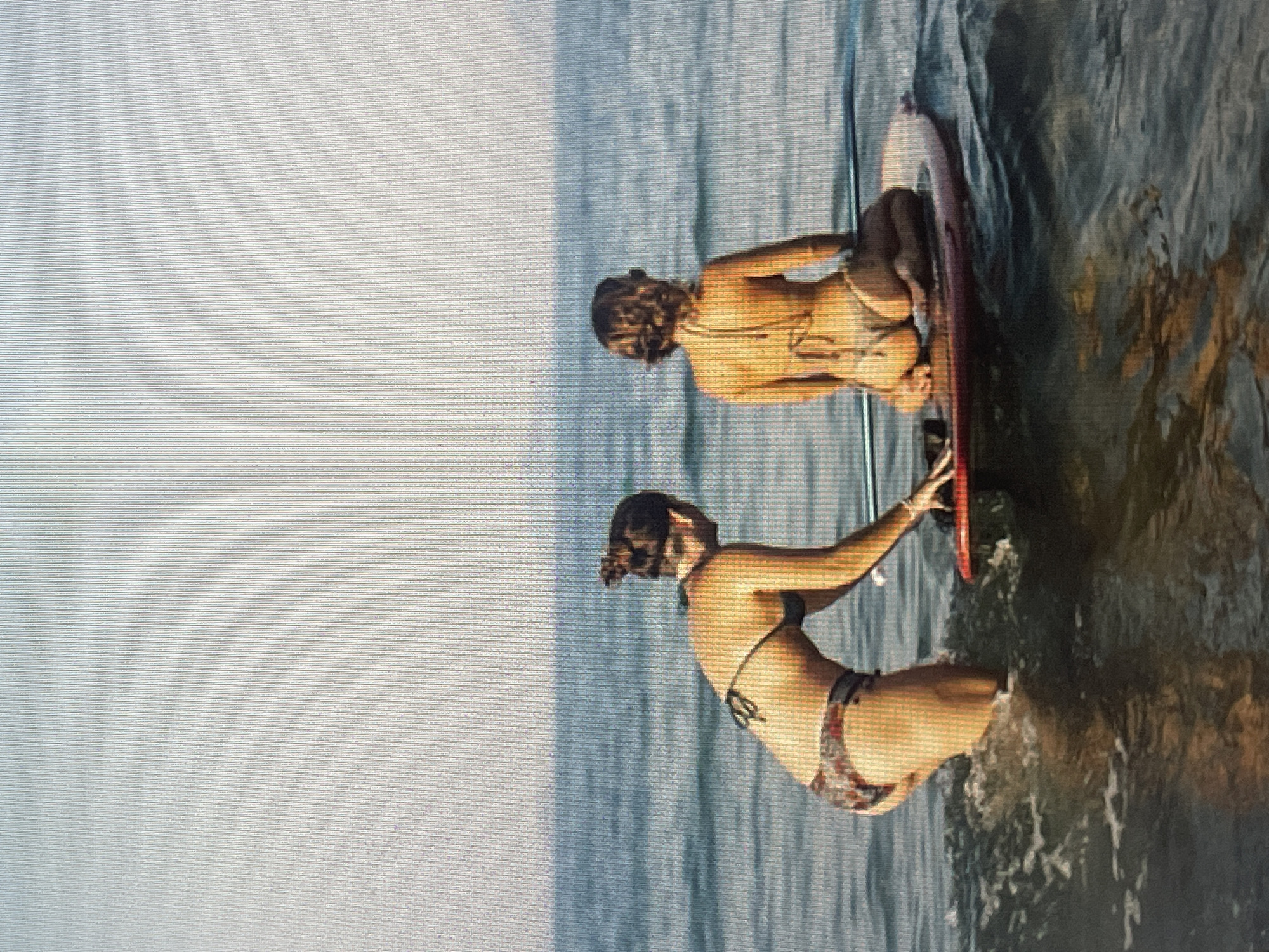 Two individuals are in the water with a surfboard. One person is kneeling on the surfboard, facing away from the camera. The other person is also kneeling on the surfboard, facing towards the camera. The water appears calm, and the lighting suggests it is daytime. The image is shot from a slightly elevated angle, looking down at the individuals and the surfboard. There is no visible text in the image.