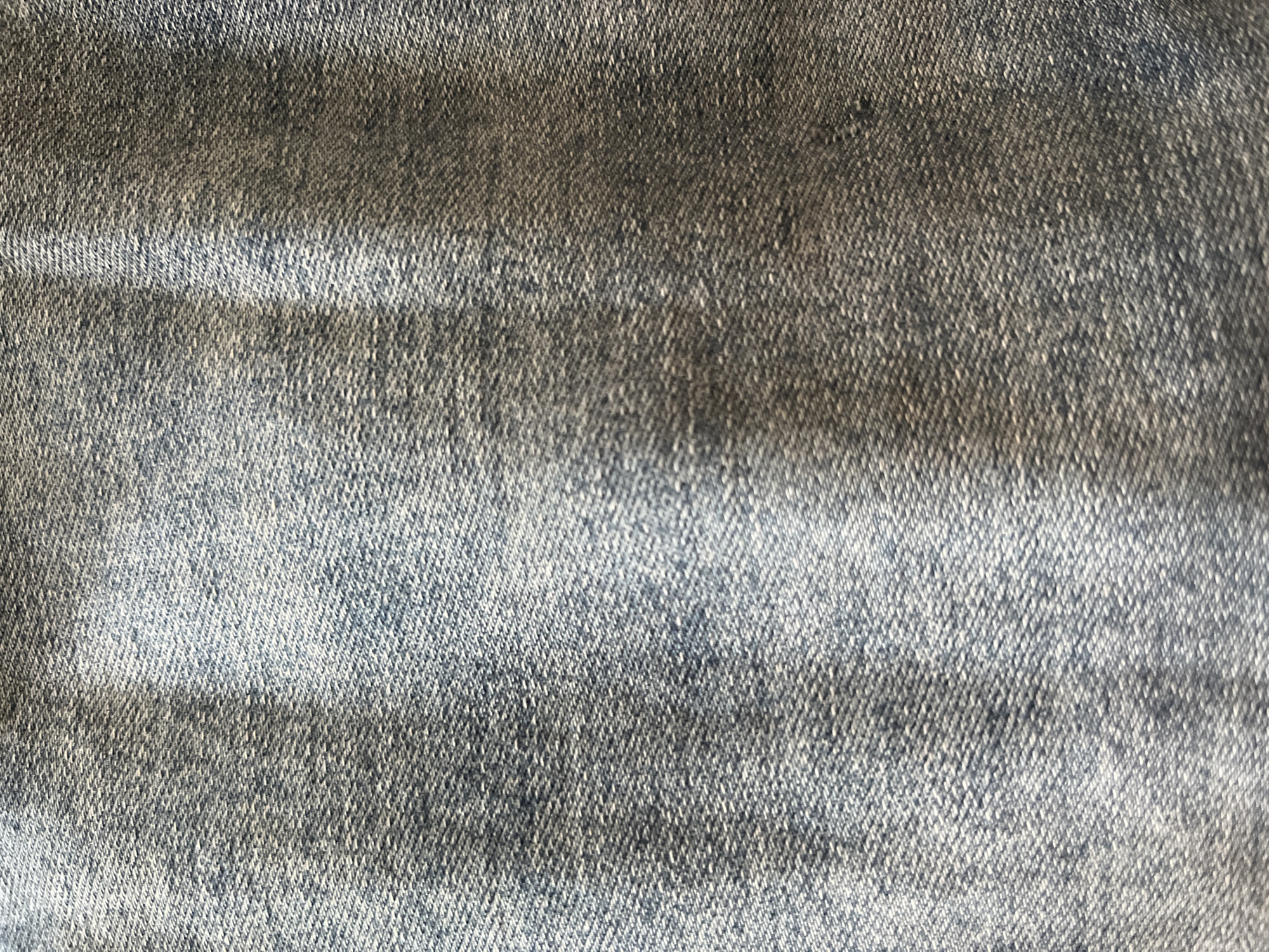 The image is a close-up, top-down view of a blue denim fabric. The texture of the denim is visible, with the characteristic diagonal weave of twill fabric. There are subtle variations in the color of the denim, with some areas appearing lighter and others darker, suggesting a slightly faded or washed look. The lighting creates some shadows, highlighting the texture and any creases or folds in the fabric. There are no identifiable objects, people, or environmental features present in the image, as it is solely focused on the textile.
