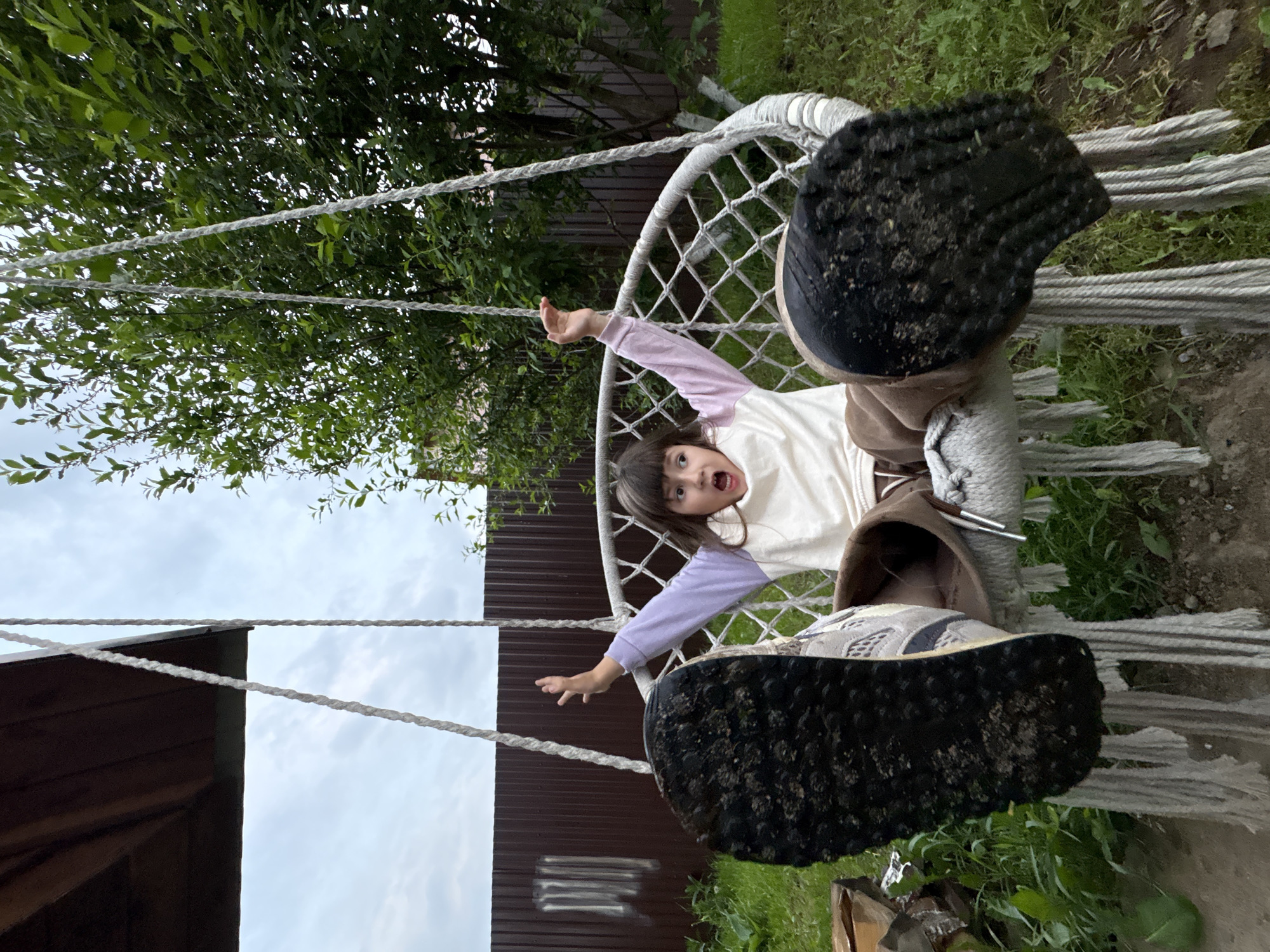 A young child wearing a light-colored long-sleeved shirt and brown shoes is in a hammock chair suspended outside, presumably in Прикамский, Russia. The soles of the child's shoes are prominently displayed towards the viewer. The child's mouth is open, and their arms are outstretched. Behind the hammock, a portion of a building with a dark-colored roof and a fence can be seen. Green foliage is visible in the background and on the ground. The sky is overcast.
