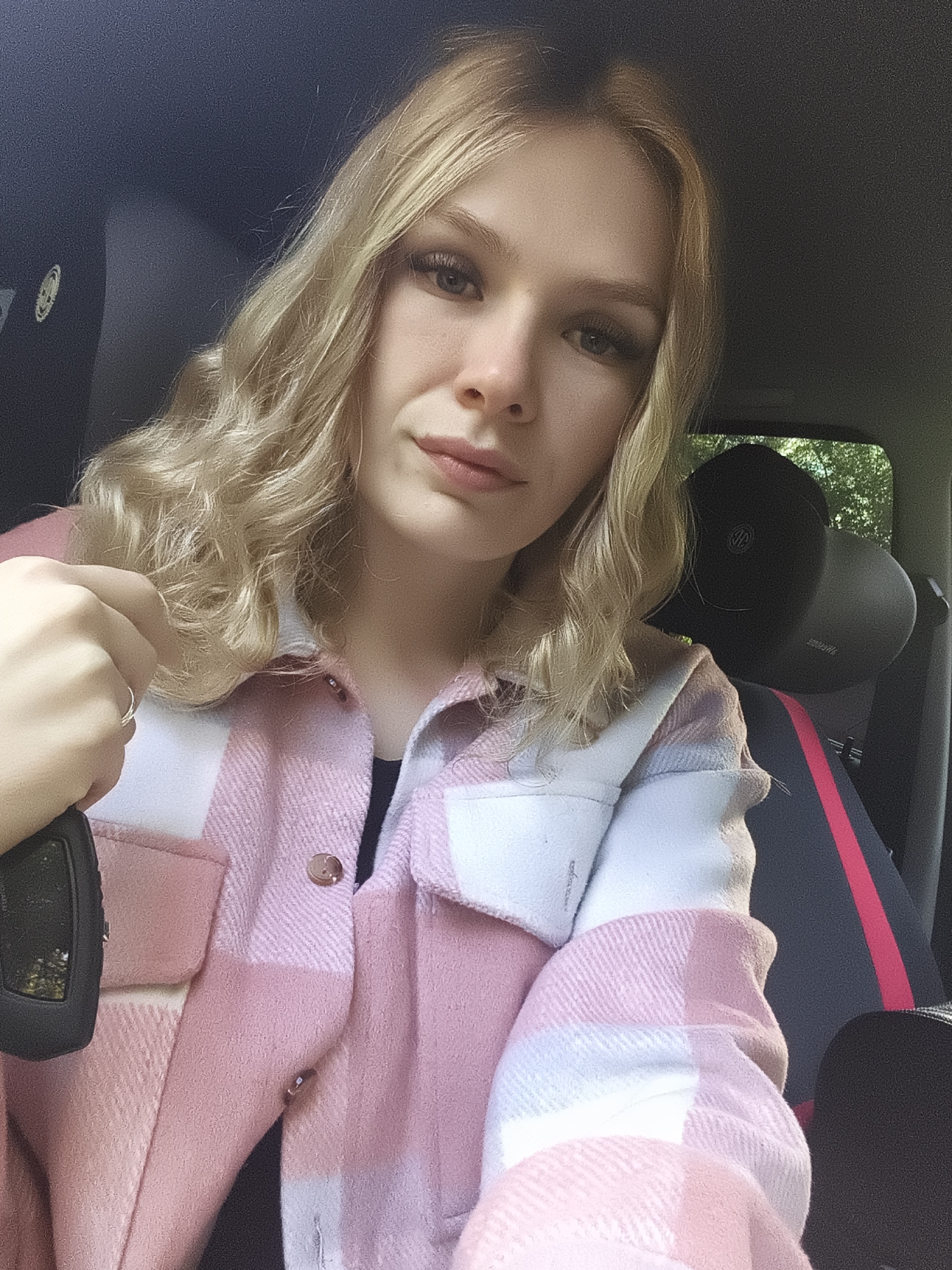 A young blonde woman is captured in a selfie-style shot, seated inside a car. She has wavy, light blonde hair that falls around her shoulders and a light complexion, with visible makeup including eyeliner and lip color. She is looking directly at the camera with a neutral to slightly soft expression.
She is wearing a cozy-looking pink and white plaid shacket or jacket over a dark top. Her left hand is visible in the lower left of the frame, holding a black car key fob, and she wears a simple silver ring on her left ring finger.
The car's interior features a dark headliner and dark seats, with a notable red stripe accenting the side of the seat visible to her right. A black headrest behind her displays a faint, circular metallic logo, resembling a car emblem (possibly Volkswagen, which is also faintly discernible as mirrored, upside-down text on the seat fabric below the headrest in some crops). Through the car windows, green foliage and trees are visible, suggesting it is daytime and the car is parked outdoors. The overall lighting is bright and natural. The provided context indicates the location is Izhevsk, Russia.