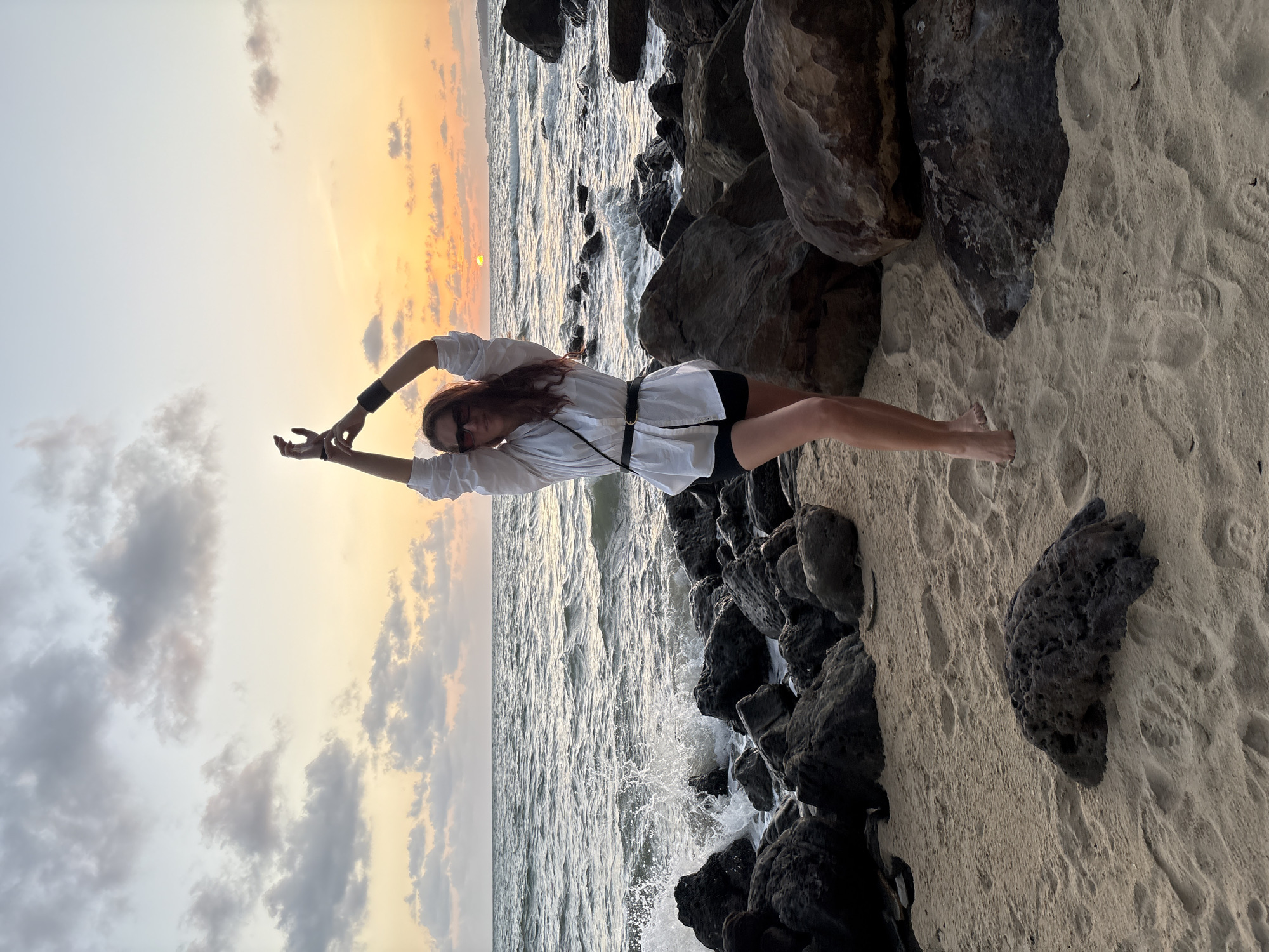 A woman stands on a sandy beach with her arms raised, silhouetted against a sunset over the ocean. She is wearing a white oversized shirt, black shorts, and sunglasses. The beach is rocky, with large dark rocks scattered along the shore. Waves crash against the rocks, creating white foam. The sky is filled with dramatic clouds in shades of orange, yellow, and gray. The overall impression is one of a serene and slightly dramatic natural scene.