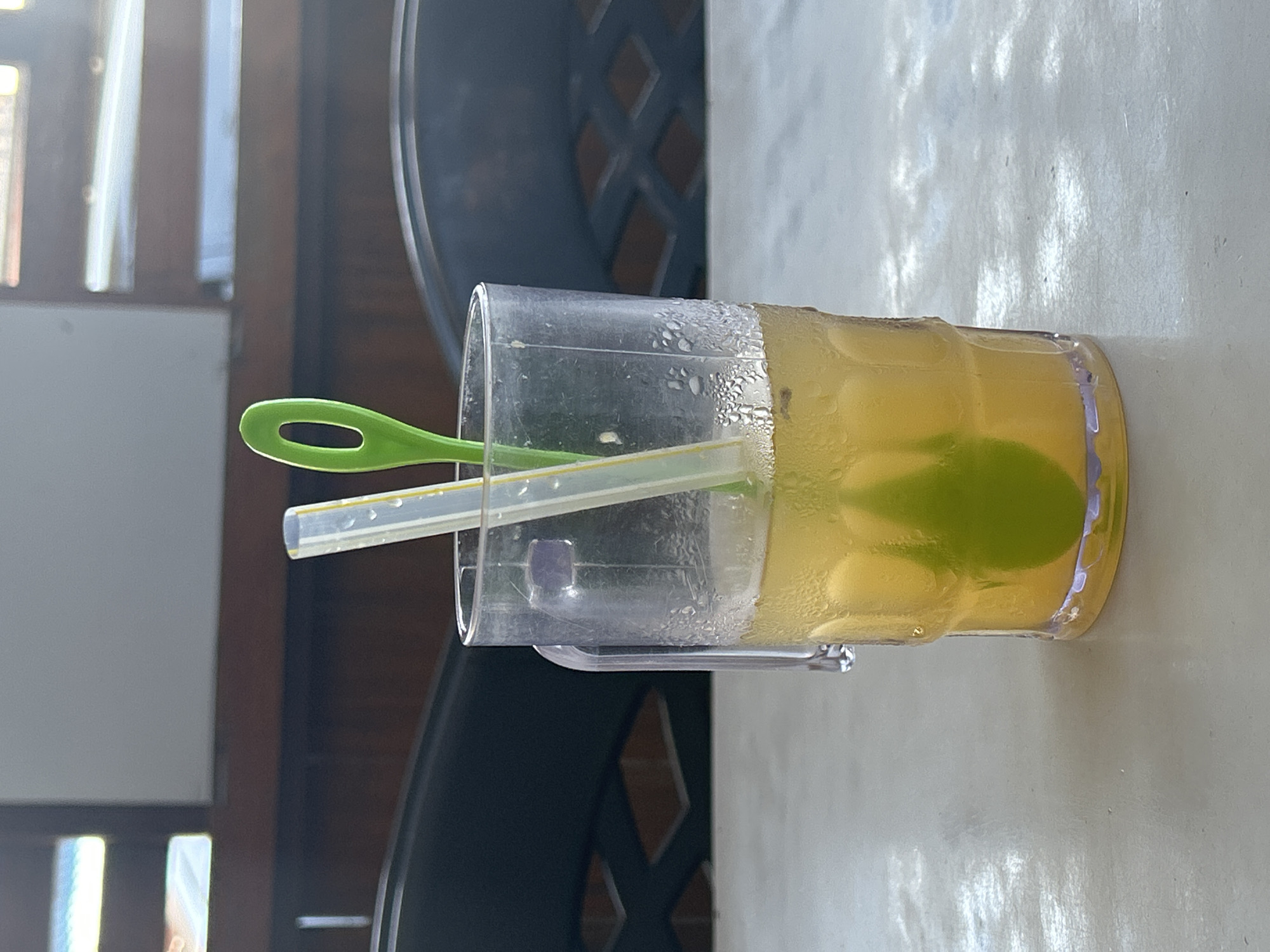 A clear, ribbed glass mug is filled with a light yellow, cloudy beverage. Inside the mug, a green lime slice is visible, submerged in the liquid. Two straws extend from the top of the mug: one is white with yellow accents and has water droplets on it, and the other is bright green and appears to be a spoon-straw. The mug rests on a light-colored, textured surface, possibly a table. In the background, dark chairs are partially visible, and behind them, a wooden wall with horizontal slats and what appears to be a window with horizontal blinds is present. The lighting suggests an indoor setting, potentially a restaurant or cafe in Mandaong, Indonesia.