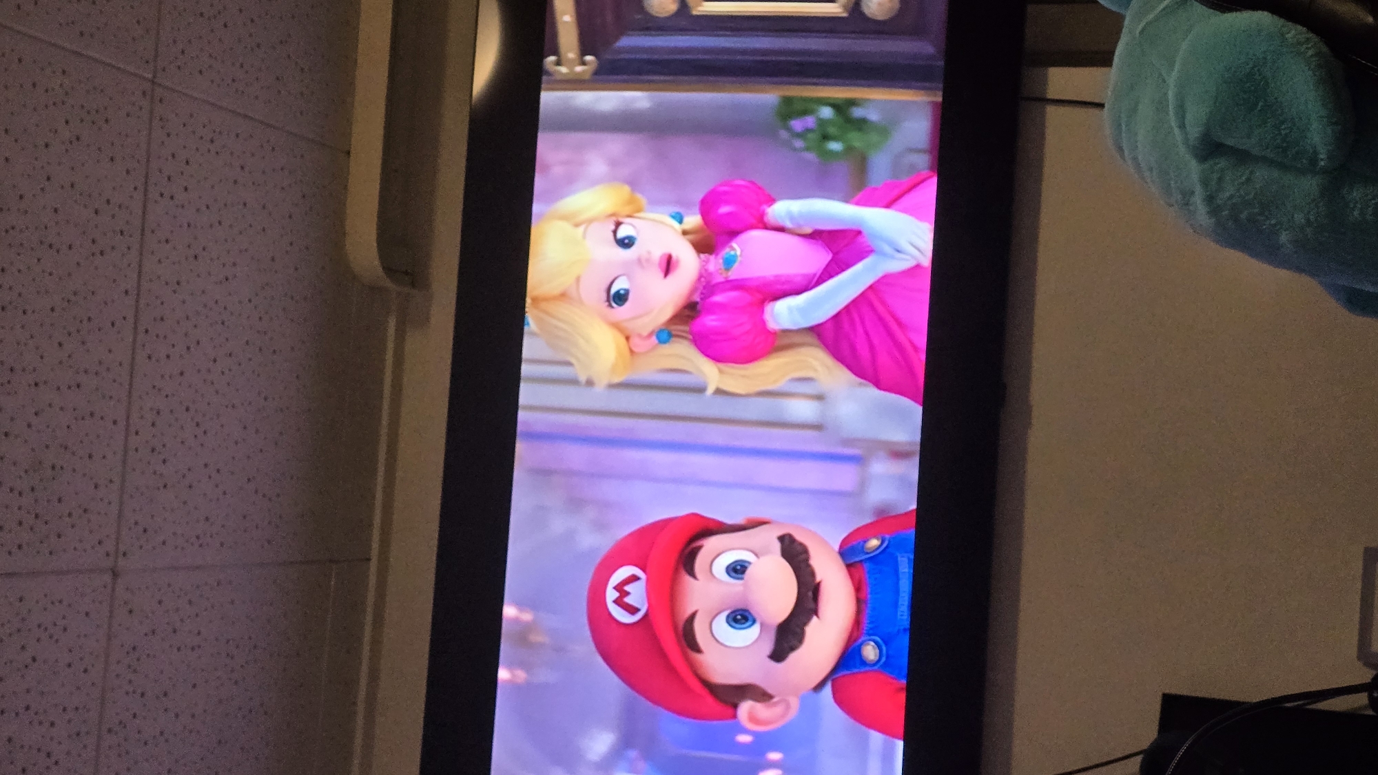 The image shows a television screen displaying characters Mario and Princess Peach. The environment includes a wall with a textured surface, what appears to be an air vent, and a dark object possibly a plush doll. The location is unspecified but is assumed to be in Zirndorf, Germany.