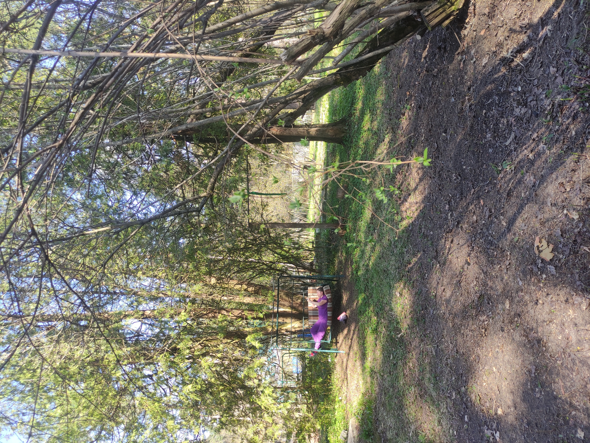 The image presents an outdoor scene, possibly a park or wooded area, with a man reclining on a swing. The individual is wearing a purple outfit. The swing appears to be constructed from metal and has a cushioned seat. The area is shaded by trees, which are a significant presence in the frame. The ground is covered with grass and dirt, indicating a natural setting. A closer inspection of the image suggests the scene is situated in a rural setting, further supported by the presence of trees and foliage. There is no information available regarding the image location being Kratovo, Russia.
