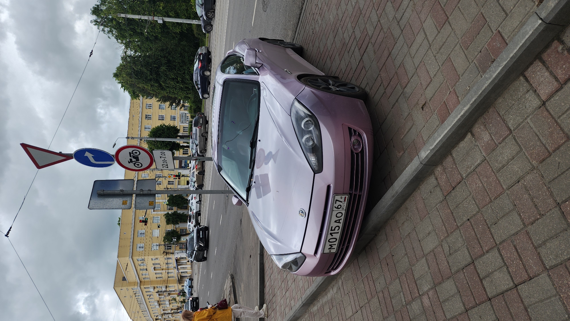 The image shows a light purple car parked on a brick sidewalk in Smolensk, Russia. The car, facing forward, has a license plate reading "M015 AO 67 RUS." The sidewalk is lined with a gray concrete curb. To the left of the car is a street with multiple parked cars and a series of traffic signs including yield, mandatory turn, and no motorcycle from 22:00 to 7:00. A person wearing a yellow jacket and light-colored pants is standing near the front of the car. In the background, there is a large yellow building with rows of windows, and trees are visible. The sky is overcast.