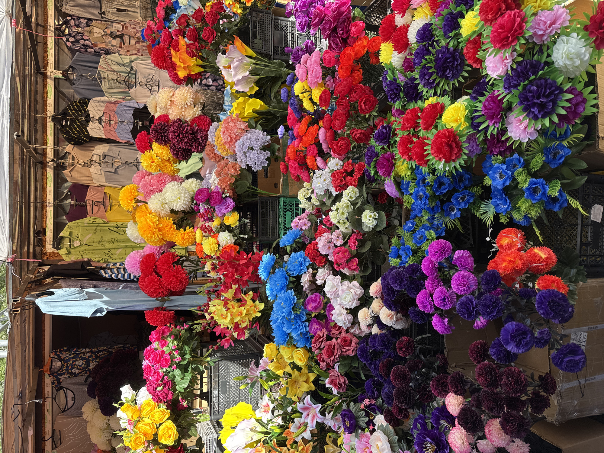 The image depicts a market stall in an unidentified city. The stall displays a variety of colorful artificial flowers arranged in clusters, interspersed with black plastic crates. To the left, clothing items hang on a rack. The stall is partially covered by a white and brown canopy. The light appears bright, suggesting it is daytime.