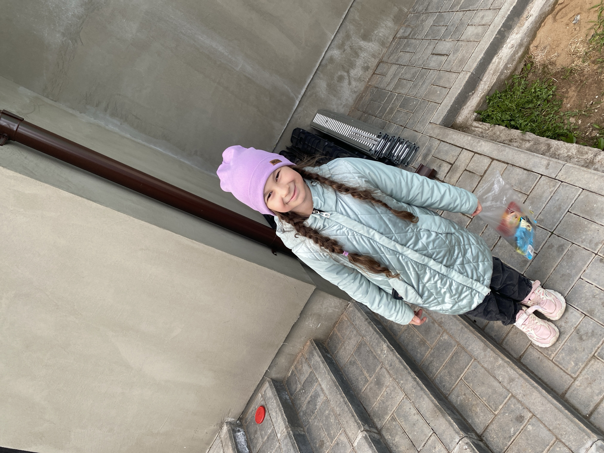 A young girl with braided hair, wearing a lilac hat, light blue quilted jacket, dark pants, and pink sneakers, stands on a grey brick patio. She holds a clear plastic bag with colorful contents. Behind her is a building with a brown rain gutter and grey walls. To her right is a patch of greenery. The location is Izhevsk, Russia.