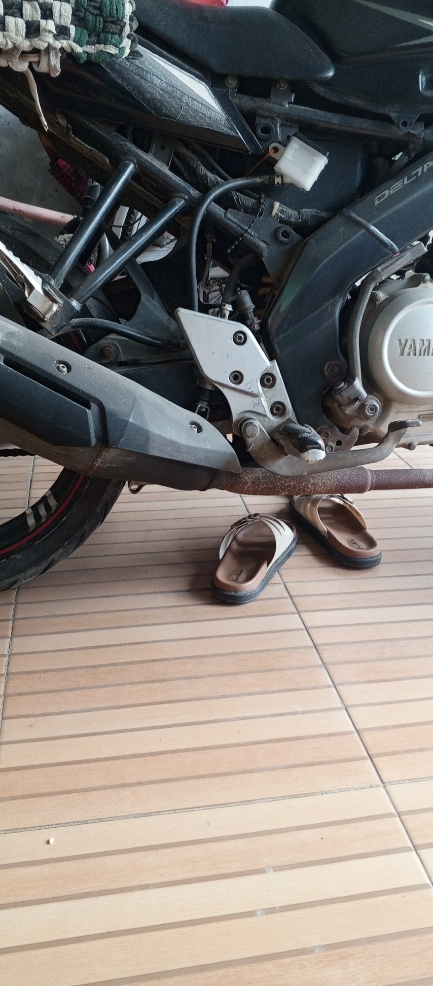 The image shows a close-up view of the lower section of a motorcycle, specifically focusing on the exhaust pipe, footpeg assembly, and a small white reservoir tank, likely for brake fluid. Two slip-on sandals, one white and beige with a patterned strap, and the other beige with a woven strap, are placed on the tiled floor beneath the motorcycle. The floor appears to be made of light brown rectangular tiles with darker grout lines. The motorcycle is positioned outdoors, as indicated by the tiled surface and the presence of shadows. The overall scene suggests a casual, perhaps domestic setting. The location is specified as Karawang, Indonesia.