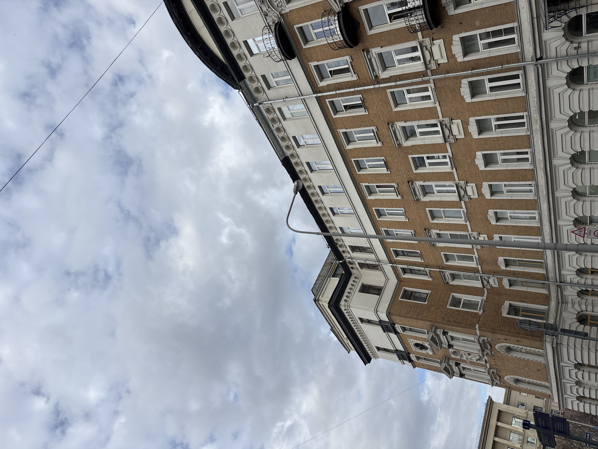 The image depicts buildings in Moscow, Russia. The buildings are multi-story with windows, architectural details, and a combination of brick and white facades. The sky is cloudy. Street lighting is visible.