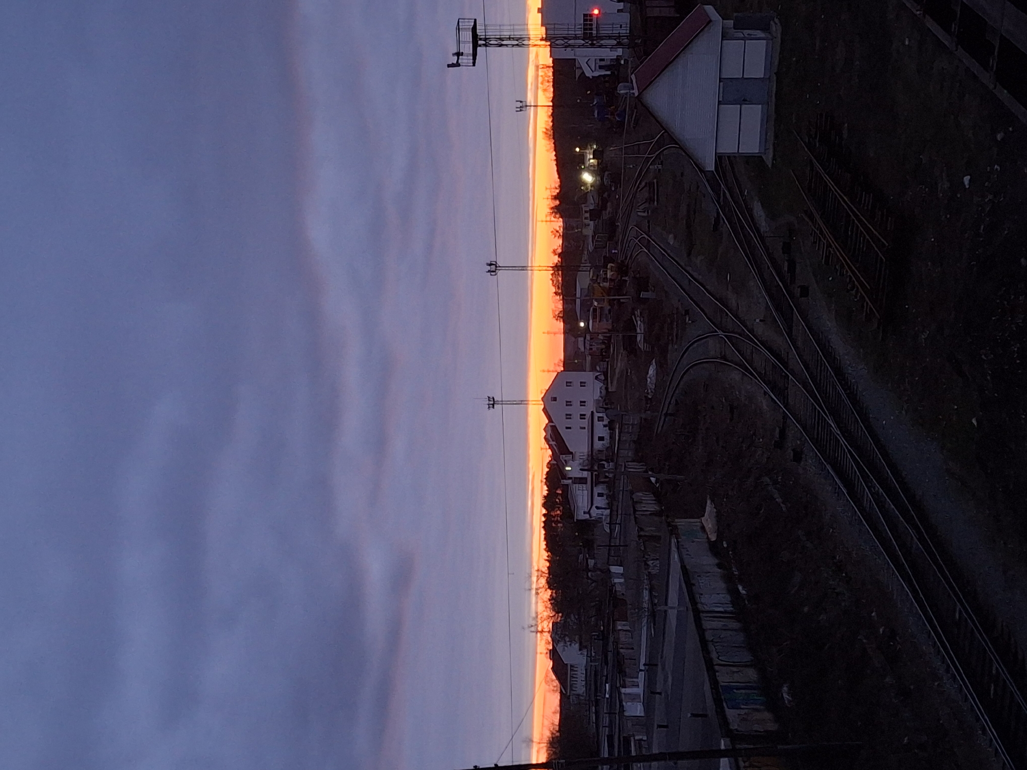 This image captures a railway or industrial area in Chaikovsky, Russia, during what appears to be sunset or sunrise. The sky dominates the upper portion of the frame, transitioning from a deep blue-purple at the top to a brilliant, intense band of orange and red along the horizon. Faint, wispy clouds are visible against the dramatic sky.
Below the colorful horizon, a dark silhouette of trees or distant vegetation stretches across the landscape. The midground and foreground reveal a complex network of railway tracks, several of which curve and converge, suggesting a shunting yard or a busy rail line. Various buildings are scattered throughout the scene; one prominent structure on the right features a red gabled roof and white walls, while another, smaller white building is visible further along. In the distance, another white building with multiple windows can be seen.
Several tall, slender metal structures, likely power line pylons or signal towers, stand silhouetted against the bright horizon. One of these structures displays a distinct red light. Artificial lights from buildings and other ground installations cast small, warm glows, contrasting with the twilight. The overall scene is devoid of visible people or specific activity, conveying a calm and serene atmosphere at the close or start of a day. No discernible text is present in the image.