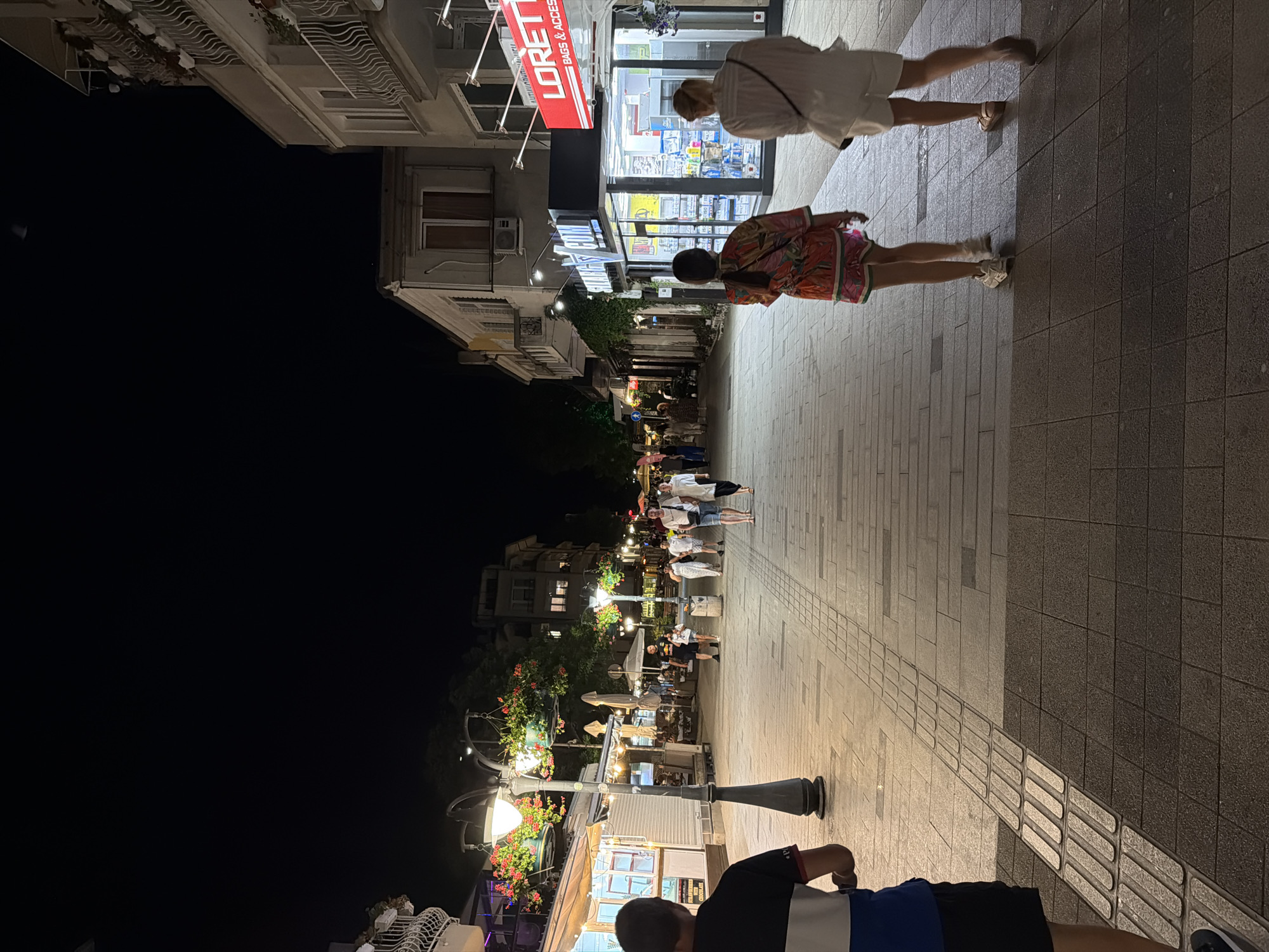 The image depicts a lively pedestrian street in Burgas, Bulgaria, at night. The street is lined with shops and restaurants, illuminated by streetlights and the glow from storefronts. Several people are strolling along the street, some in groups and others individually. The architecture suggests a European coastal town. The scene captures the bustling atmosphere of a summer evening, with people enjoying the outdoor ambiance.