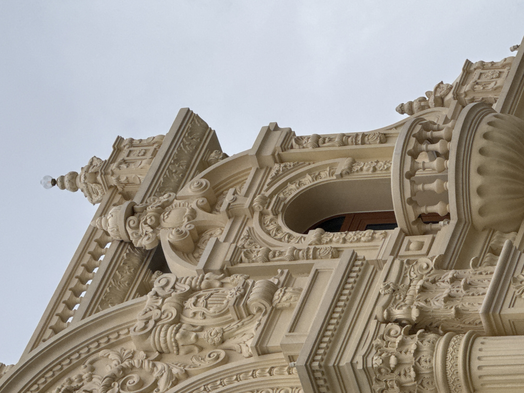 The image shows a close-up of the ornate architecture of the Da Lat Palace Hotel in Da Lat, Vietnam. The focus is on the upper facade, which features intricate carvings, balconies, and decorative elements. The building material appears to be a light-colored stone or stucco, with a creamy beige hue. The sky in the background is overcast and a pale gray. Several architectural details are visible, including Corinthian columns, arched windows, decorative moldings, and a balustrade. The overall impression is one of grandeur and classical design. There are no people or animals visible in the image. The lighting suggests it is daytime, possibly late afternoon or early morning given the diffused light.