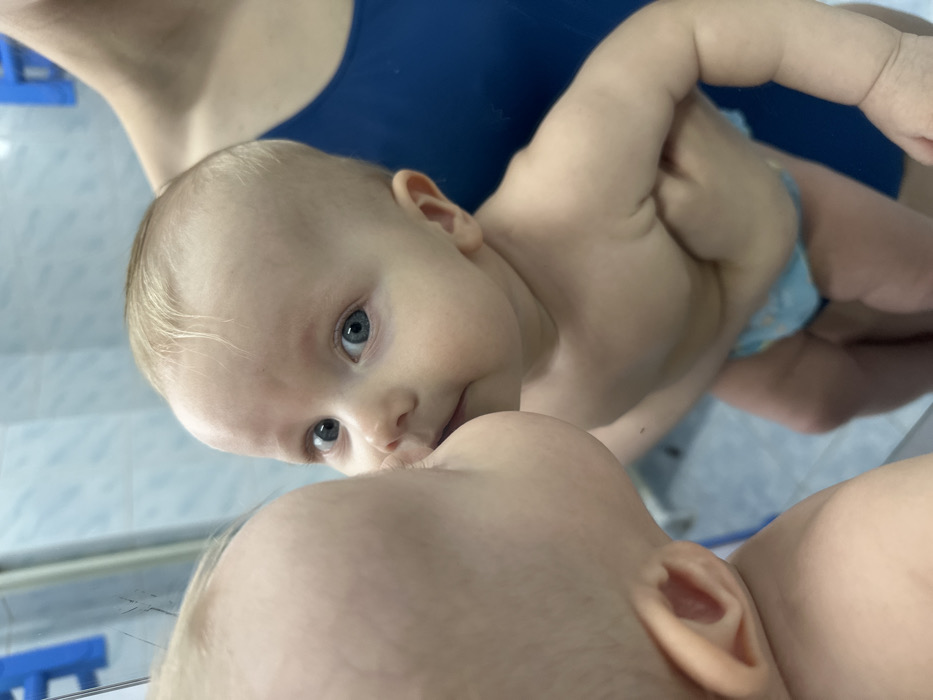 The image depicts an indoor scene featuring an infant being held by an adult in front of a mirror. The specific location within Orekhovo-Zuyevo, Russia, is not discernible from the visual cues; the background suggests a residential interior, possibly a bathroom, with a light blue wall.
A light-skinned infant, with sparse light blonde hair and blue eyes, is positioned facing the mirror. The infant is wearing a light blue diaper with a visible pattern. The infant's upper torso and arms are bare. Its face is directed towards the mirror, with its mouth and lower face pressed against or in very close proximity to its reflection. The infant's gaze is directed forward, seemingly towards the viewer.
A partially visible adult supports the infant. Only the adult's upper chest, shoulder, and arms are visible. The adult is wearing a dark blue tank top or similar garment. Their hands are positioned to hold and stabilize the infant around its torso.
The mirror surface, occupying the left side of the frame, reflects the infant's profile. The background behind the infant and adult is a uniform light blue wall, possibly tiled. A partial blue object, possibly a handle or fixture, is visible in the upper left background.