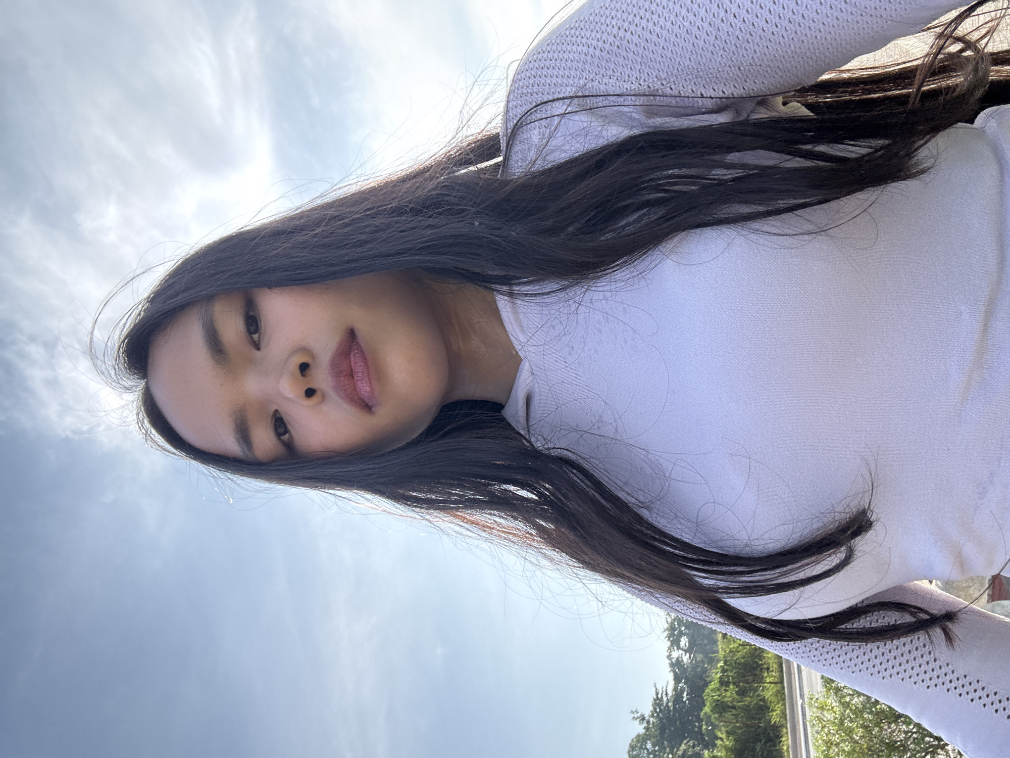 The image is a selfie taken outdoors, with the subject facing the camera. The subject is a young female with long, dark hair and fair skin. She is wearing a white hooded top with a textured pattern on the sleeves. The background consists of a bright blue sky with some clouds, and a hint of greenery and a road in the lower left corner, suggesting an outdoor urban or suburban setting. There are no other individuals or specific landmarks that definitively place the location within Davao City, Philippines, though the lighting and vegetation are consistent with a tropical climate.