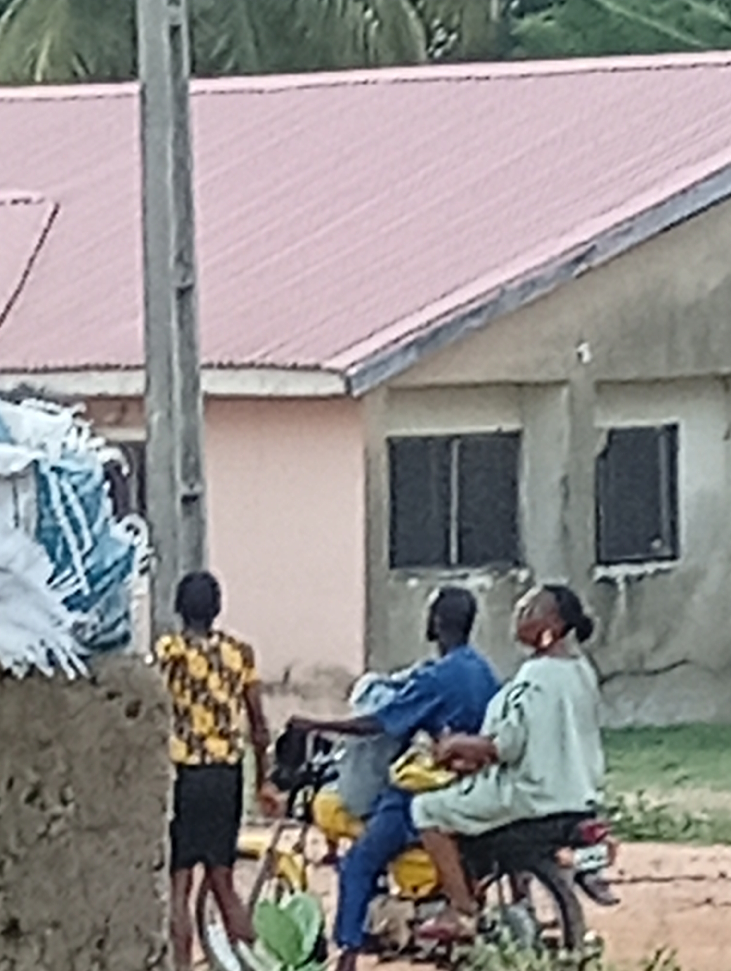 In Adankolo, Nigeria, the image captures three individuals in an outdoor setting near buildings. One person stands on the left in a patterned yellow shirt and black shorts. Two other people are on a yellow motorcycle, the driver in a blue shirt and pants, and the passenger in a light-colored top. The buildings behind them have a pink roof and dark-framed windows. A utility pole is visible near the left building. There's green vegetation in the foreground.
