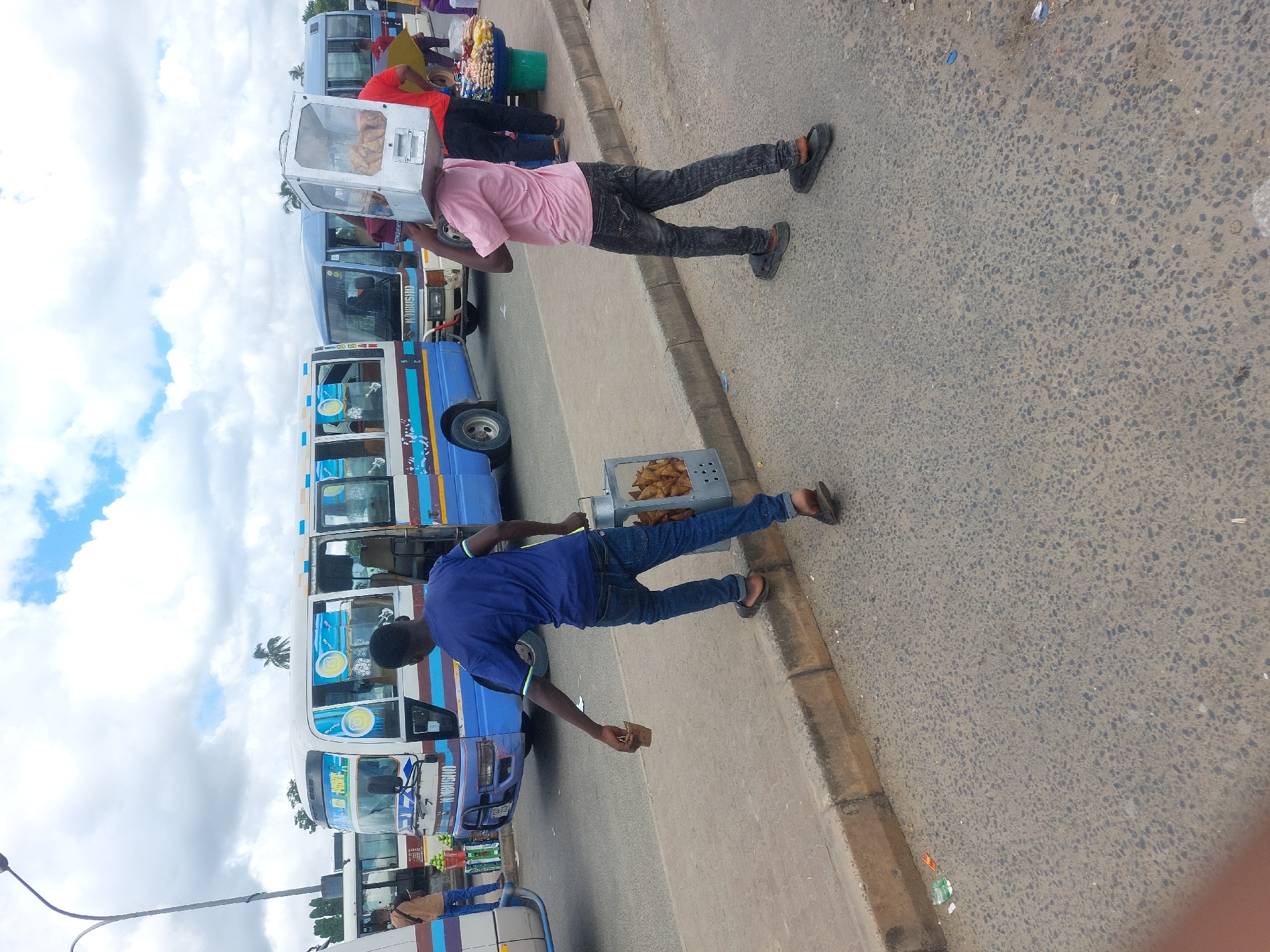 The image shows a street scene in Dar es-Salaam, Tanzania. Two men are selling food near a row of blue and white minibuses. One man wears a pink shirt and carries a glass display case on his head filled with baked goods. The other man wears a blue shirt and carries a tray of samosas. The street is paved and shows signs of wear. The sky is cloudy.