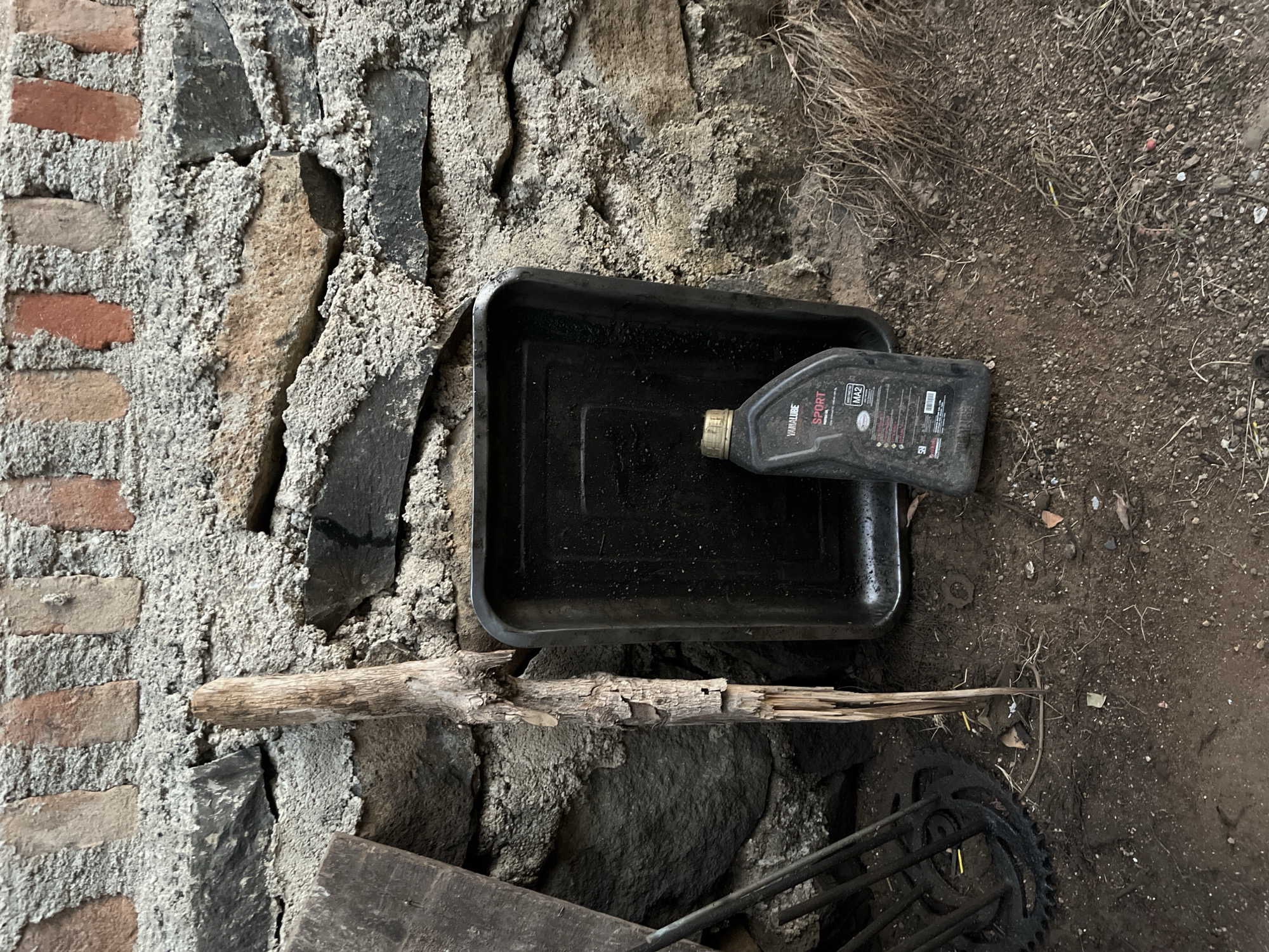 An oil change station is depicted in front of a stone wall in Kebumen, Indonesia. A black plastic oil drain pan sits on the ground, with a black plastic bottle of Yamalube Sport oil placed to its right. A weathered wooden stick stands vertically between the pan and the wall, and to the left, a motorcycle chain and sprocket are visible. The ground is covered in dirt and debris.