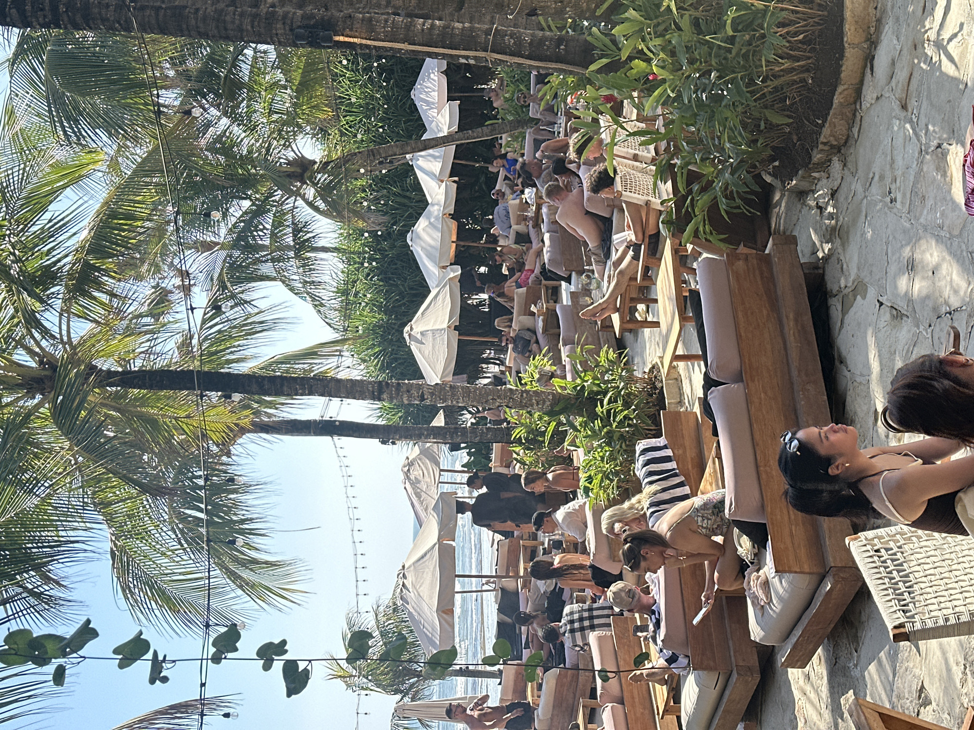 The image depicts a crowded outdoor seating area at a beachside establishment in North Kuta, Indonesia. Numerous patrons are seen relaxing on low wooden lounge beds, many of which are equipped with cushions and small side tables. White umbrellas provide shade for some of the seating arrangements.
In the foreground, several individuals are seated or reclining. A woman in the immediate foreground, wearing a dark swimsuit and sunglasses, looks to her right. Behind her, partially visible, is another person. To the left and further back, a woman in a patterned swimsuit is seated, holding a mobile phone.
Further into the scene, the beach and ocean are visible in the background. Several palm trees are interspersed throughout the area, and lush green foliage lines the perimeter of the seating space. String lights are visible hanging between the trees. The ground is paved with irregular stone slabs.
The atmosphere appears relaxed, with people socializing and enjoying the beachfront environment. The lighting suggests it is daytime, with bright sunlight casting shadows.