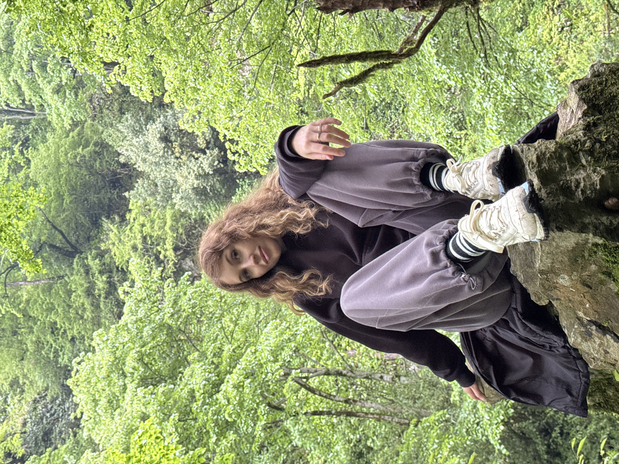A young woman with curly brown hair is sitting cross-legged on a rocky outcrop in a forest setting. She is wearing gray sweatpants with black and white striped socks and white sneakers, and a dark-colored sweatshirt. She has rings on her fingers. The background consists of lush green foliage and trees, suggesting a natural environment. The image was taken in the city of Tsabal, Abkhazia.