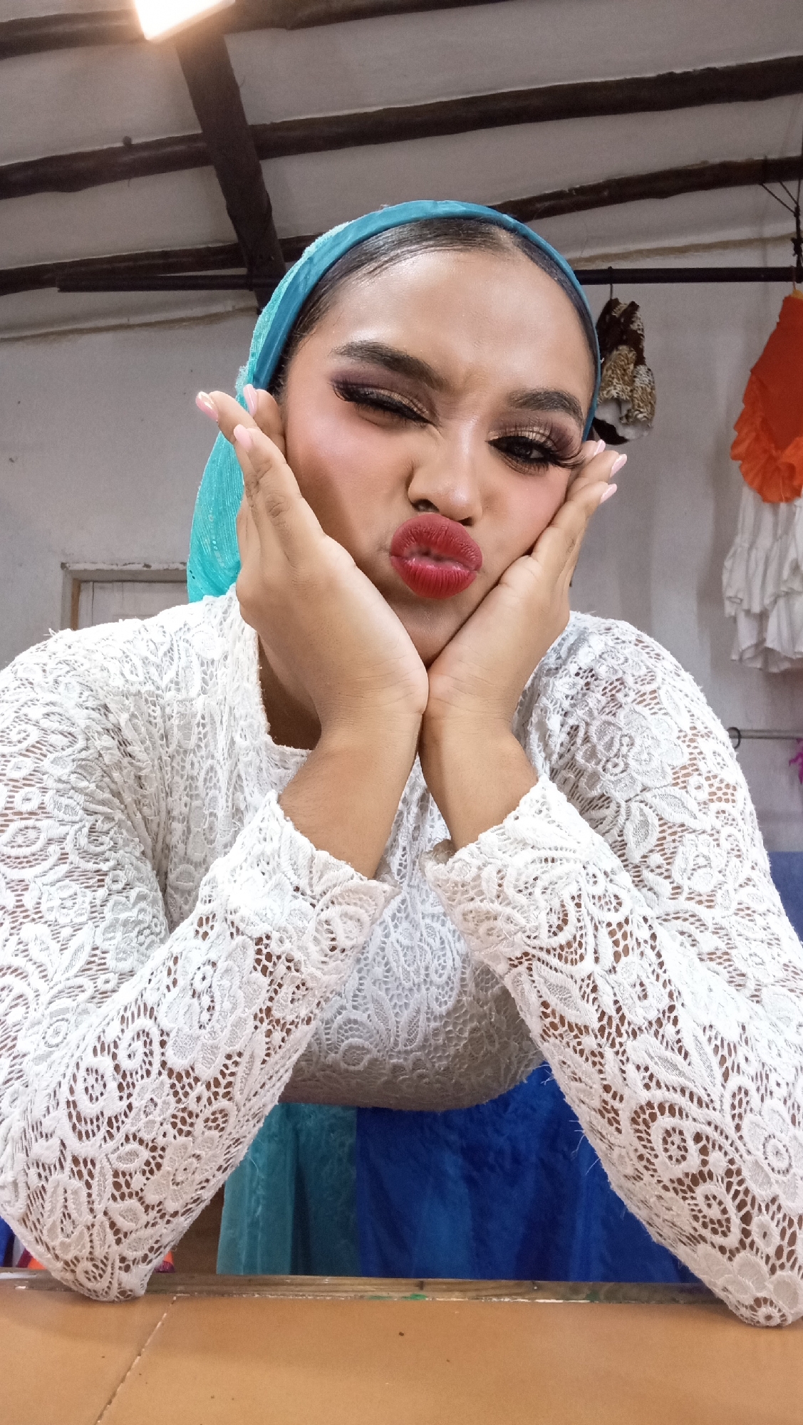 A young individual, appearing female, with medium skin tone and dark hair styled under a turquoise fabric headband, is depicted indoors in Porlamar, Venezuela. The subject is wearing a white long-sleeved lace blouse and a blue garment partially visible below. Their face is made up with red lipstick, dark eye makeup, and false eyelashes. The individual is striking a playful pose, making a "duck face" with pursed red lips and partially squinted eyes, while cupping their cheeks with both hands, which display light pink nail polish. The background features a light-colored ceiling with dark exposed wooden beams and a bright rectangular light fixture in the upper left. To the right, various items of clothing, including an orange ruffled fabric, a white ruffled garment, and a patterned garment, hang on hooks. A light-colored wall with a dark rectangular feature is visible on the left. The foreground consists of a light brown, flat surface, possibly a table or box.