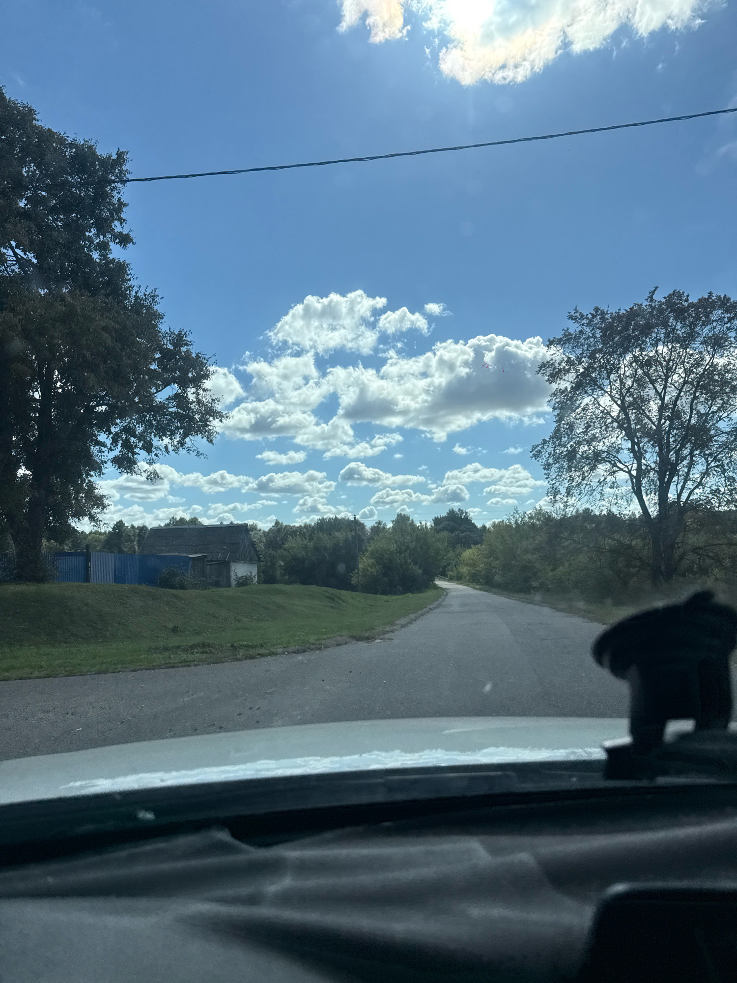 The image shows a view from inside a vehicle looking out onto a paved road that curves to the left, leading into a rural landscape under a bright blue sky with scattered white clouds. On the left side of the road, there is a grassy embankment with a blue fence and a small, aged building with a dark roof. Two large trees frame the scene, one on the left and another on the right. The dashboard of the vehicle is visible in the foreground, with a dark, shaped object, possibly a car accessory, mounted on it. The sky is lit by the sun, which is partially obscured by a cloud. The overall impression is a sunny day in a rural setting.