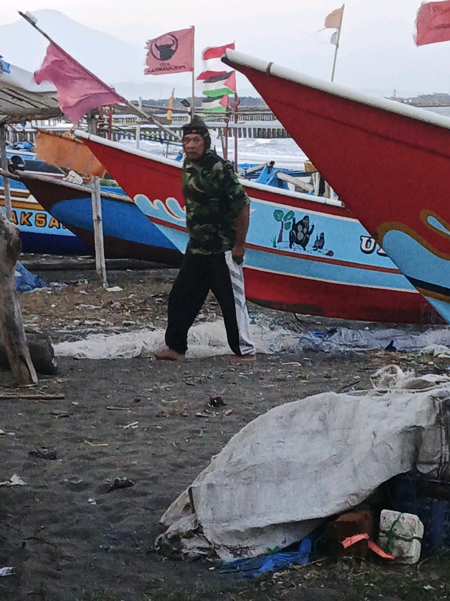 The image depicts a man standing on a beach near several boats. The man is wearing a camouflage jacket and black pants. He is barefoot. In the background, there are several fishing boats with colorful hulls and flags. One boat has a red hull with blue trim and a drawing of monkeys and trees on its side. Another boat has a red hull with white trim. There are also flags flying from poles, including a red flag with a bull symbol and other colorful flags. The beach appears to be covered in sand, debris, and some white plastic sheeting or material. The overall scene suggests a harbor or a boatyard in Indonesia, likely in Kelurahan Cilacap, Kecamatan Cilacap, given the context. The lighting is overcast, indicating a cloudy or hazy day.