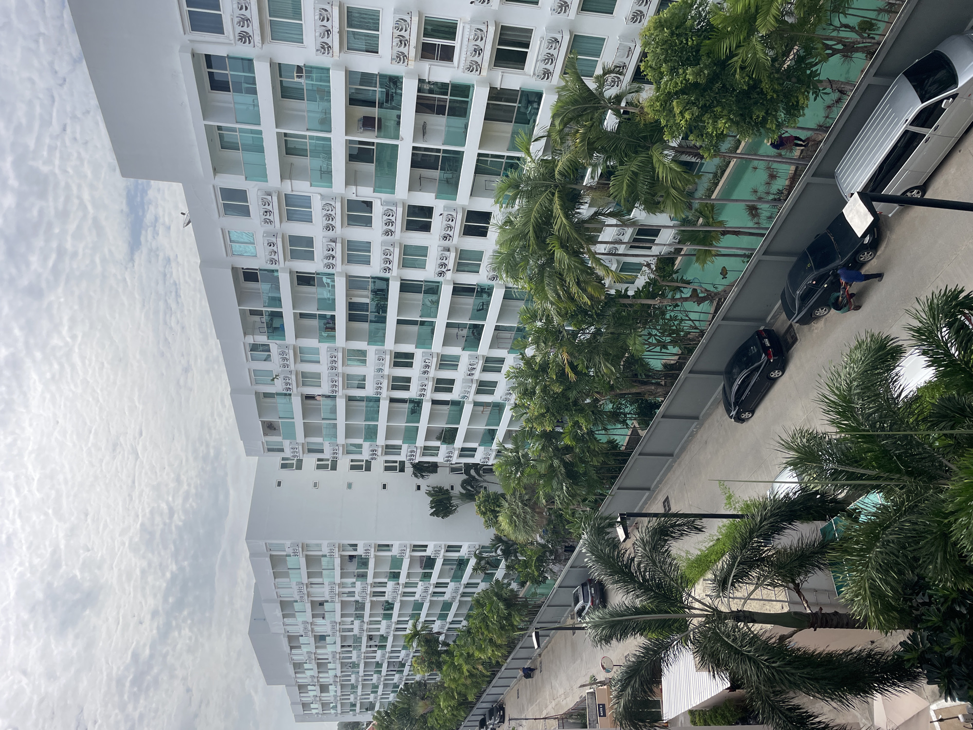 The image shows a street scene in Pattaya City, Thailand, with a long, modern white apartment complex dominating the background. The building features multiple floors with balconies, each accented by turquoise glass railings. Numerous air conditioning units are visible on the exterior of the building. In the foreground, a paved street runs alongside the complex, lined with palm trees and other lush green vegetation. Several vehicles, including black sedans and a silver minivan, are parked or driving on the street. A swimming pool with turquoise water is visible between the building and the street, also bordered by palm trees. One person is seen near the pool, and two individuals are interacting near a black car on the street. The sky is overcast with a textured cloud cover.