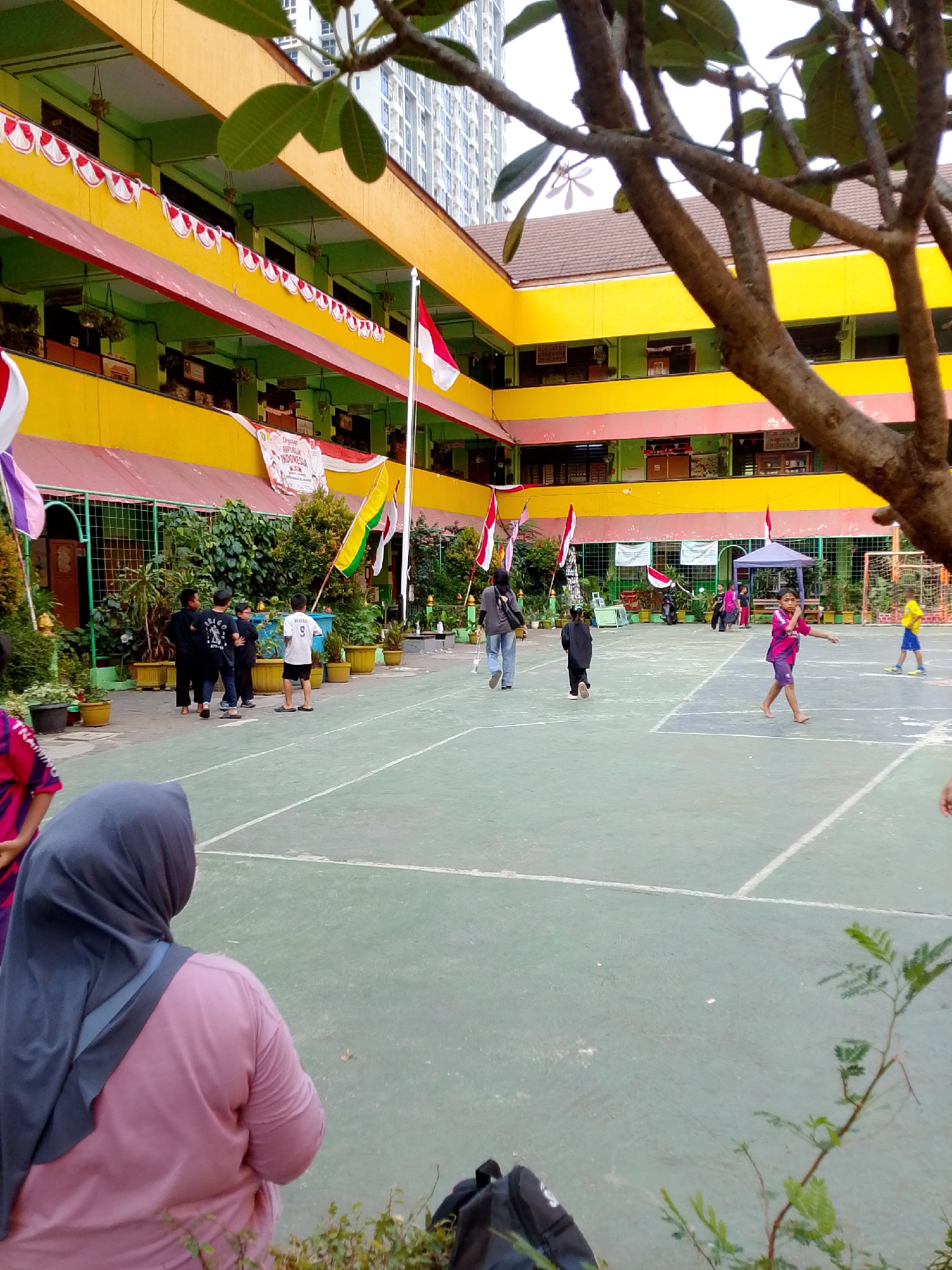In the city of Central Jakarta, Indonesia, a schoolyard hosts a group of individuals engaged in various activities. A tall building looms in the background, suggesting an urban environment. The school building itself is brightly colored with yellow and green, adorned with Indonesian flags and banners celebrating national pride. Children are seen playing on a green court, some participating in what appears to be a sports event. One child in a pink shirt and purple shorts is mid-stride, while another in a yellow shirt and blue shorts runs in the distance. A person in a pink shirt and gray hijab observes the scene from the foreground. The court is marked with white lines, indicating a designated playing area. The overall atmosphere suggests a lively school event, possibly a sports day or cultural celebration.