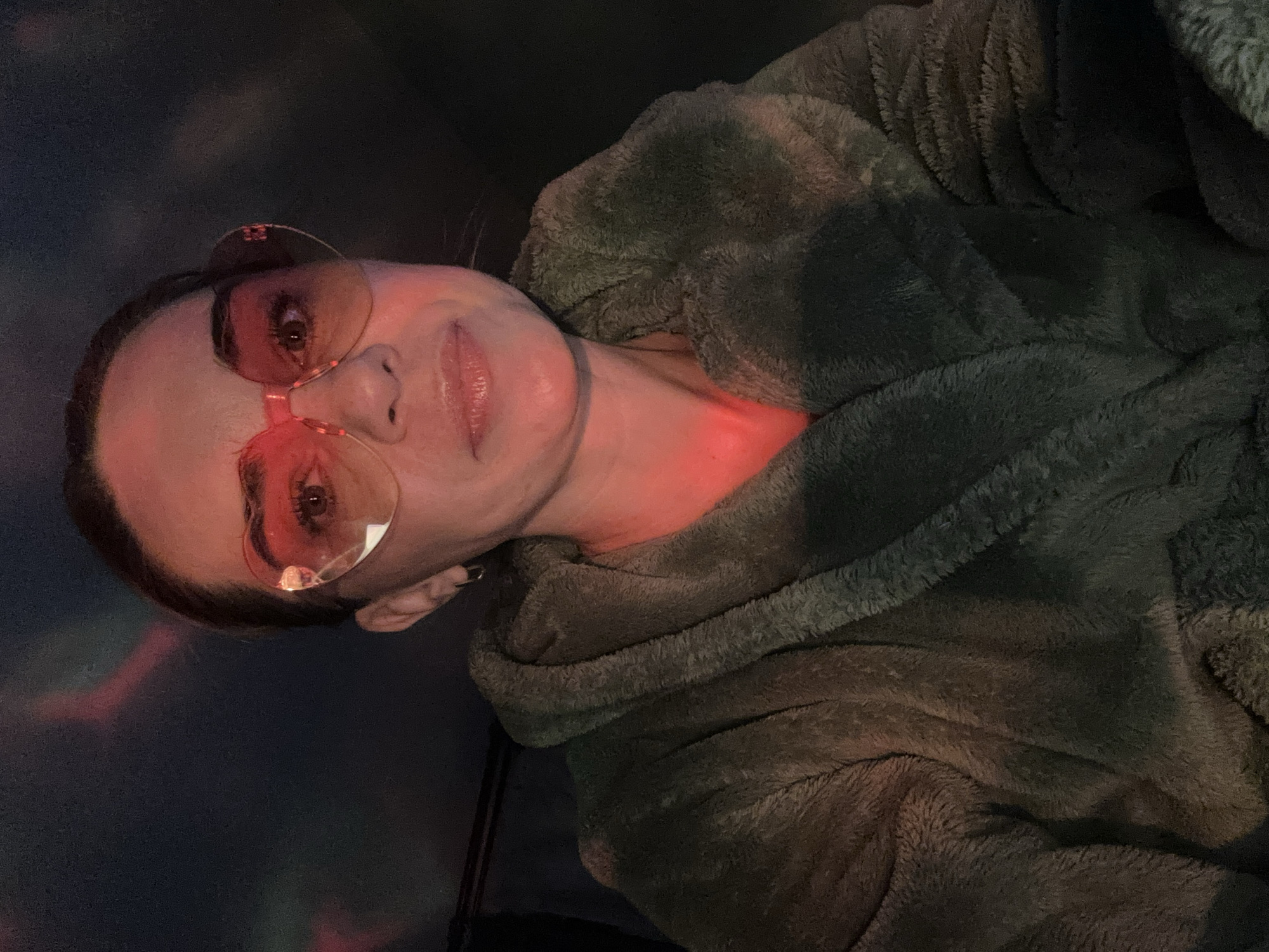 A woman is indoors wearing round, pink-tinted glasses and a fuzzy, greenish-gray robe with a hood. Her face and neck are illuminated by a red light source. The background is dark, with some faint lighting effects resembling clouds. The image is oriented vertically. There is no indication of the location being Murino, Russia.