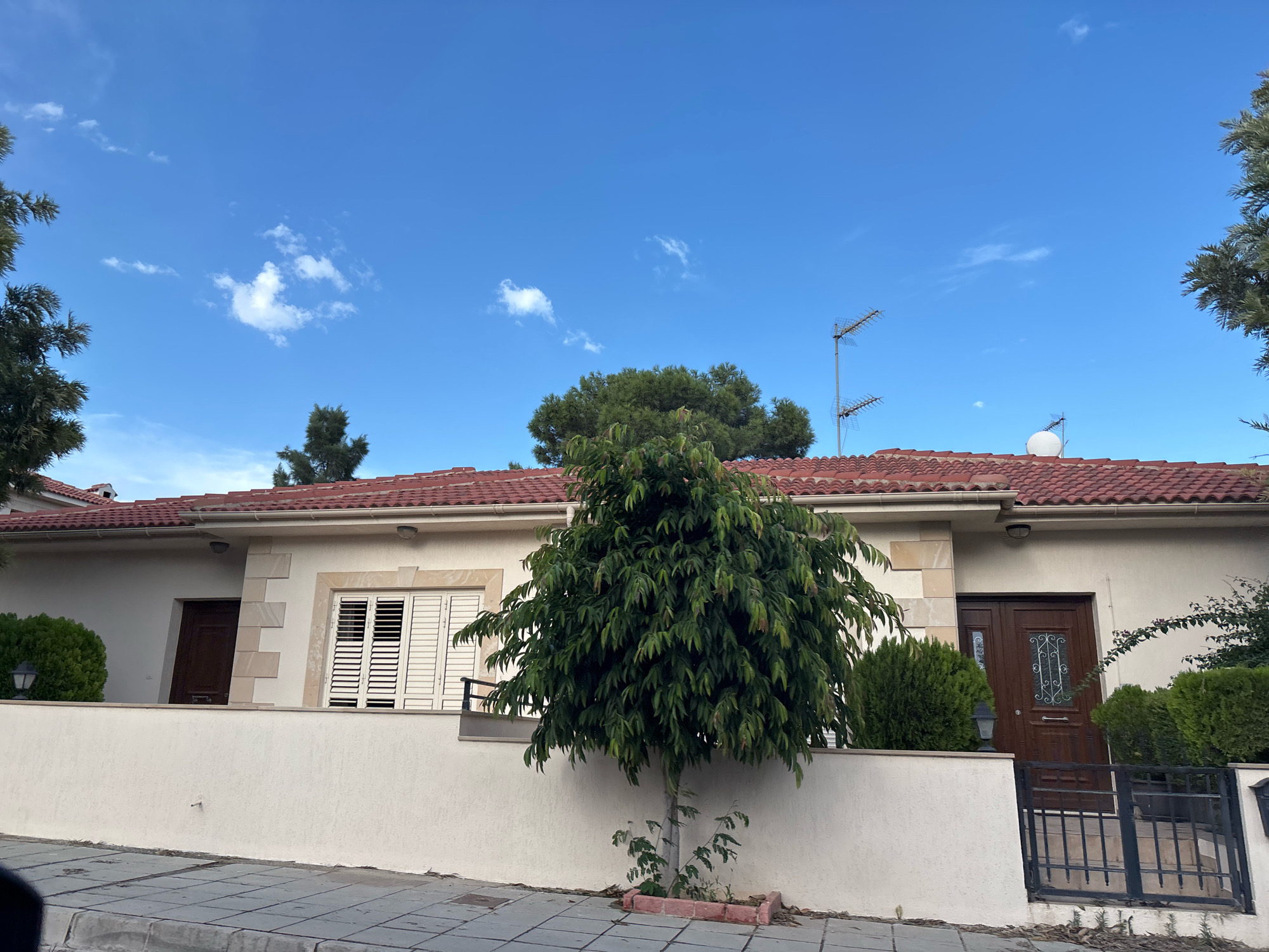 The image depicts a suburban street scene in Agios Athanasios Municipality, Cyprus. The foreground shows a paved sidewalk and a low, cream-colored wall bordering a property. Behind the wall, a single-story building with a red-tiled roof is visible. The building has light-colored walls with some stone accents. Two brown wooden doors, one with decorative glass, are visible on the facade, along with white louvered shutters on a window. A medium-sized, leafy green tree stands in front of the wall and building. To the right of the main entrance, a black metal gate with a railing leads up to the house. In the background, evergreen trees and hedges are present. The sky is blue with scattered white clouds. Two television antennas and a satellite dish are mounted on the roof.