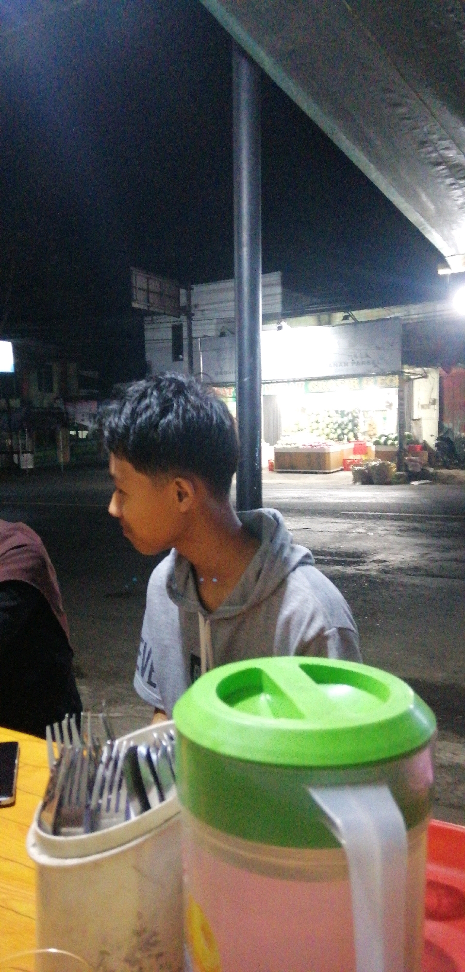 The image captures a night scene in Ajibarang, Indonesia, possibly at an outdoor food stall or eatery. In the foreground, a young person wearing a grey hooded sweatshirt is seated, looking to the left. A light blue necklace is visible on their neck. In front of them is a white container filled with cutlery, including forks and spoons, and a large pitcher with a green lid and a light-colored handle. The pitcher is pinkish-white in color with a circular yellow design.
The background shows a dimly lit street with buildings and what appears to be a market stall selling produce, with stacked fruits and vegetables visible. A tall, dark pole stands prominently near the center of the image, likely a support for a canopy or structure. The overall lighting suggests it is nighttime, with artificial lights illuminating the market area.