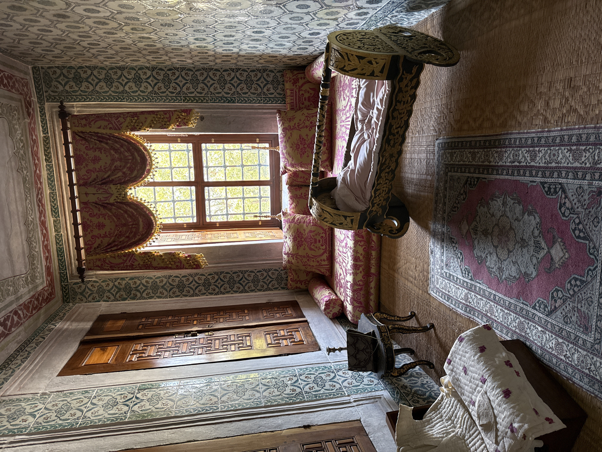 This is a photograph of a room that appears to be located in Istanbul, Turkey, likely within a historical palace or museum such as Topkapi Palace. The room is ornately decorated with patterned tiles on the walls, featuring a geometric design in green and white. The ceiling is also decorated with painted patterns in red and green.
The room contains a long, low sofa covered in a pink and gold patterned fabric, with matching bolster pillows. In front of the sofa is a wooden cradle, gilded with intricate carvings, holding a light pink bedding. To the left of the sofa, near a closed wooden door with carved panels, is a small, dark side table with ornate legs, topped with a dark decorative object that resembles a folded piece of fabric or a clutch bag, and a candlestick. Beside the side table, on the floor, is a folded white textile, possibly a quilt or blanket, with purple embroidered details, and a small white cap. A patterned rug with red, grey, and white motifs is laid out on the woven straw floor covering. The room is illuminated by natural light streaming through a window with multiple panes, covered by elaborate, draped curtains in pink and gold with fringed trim. The view through the window shows trees with green foliage. The overall impression is one of historical opulence and domestic tranquility. There are no people visible in the scene.