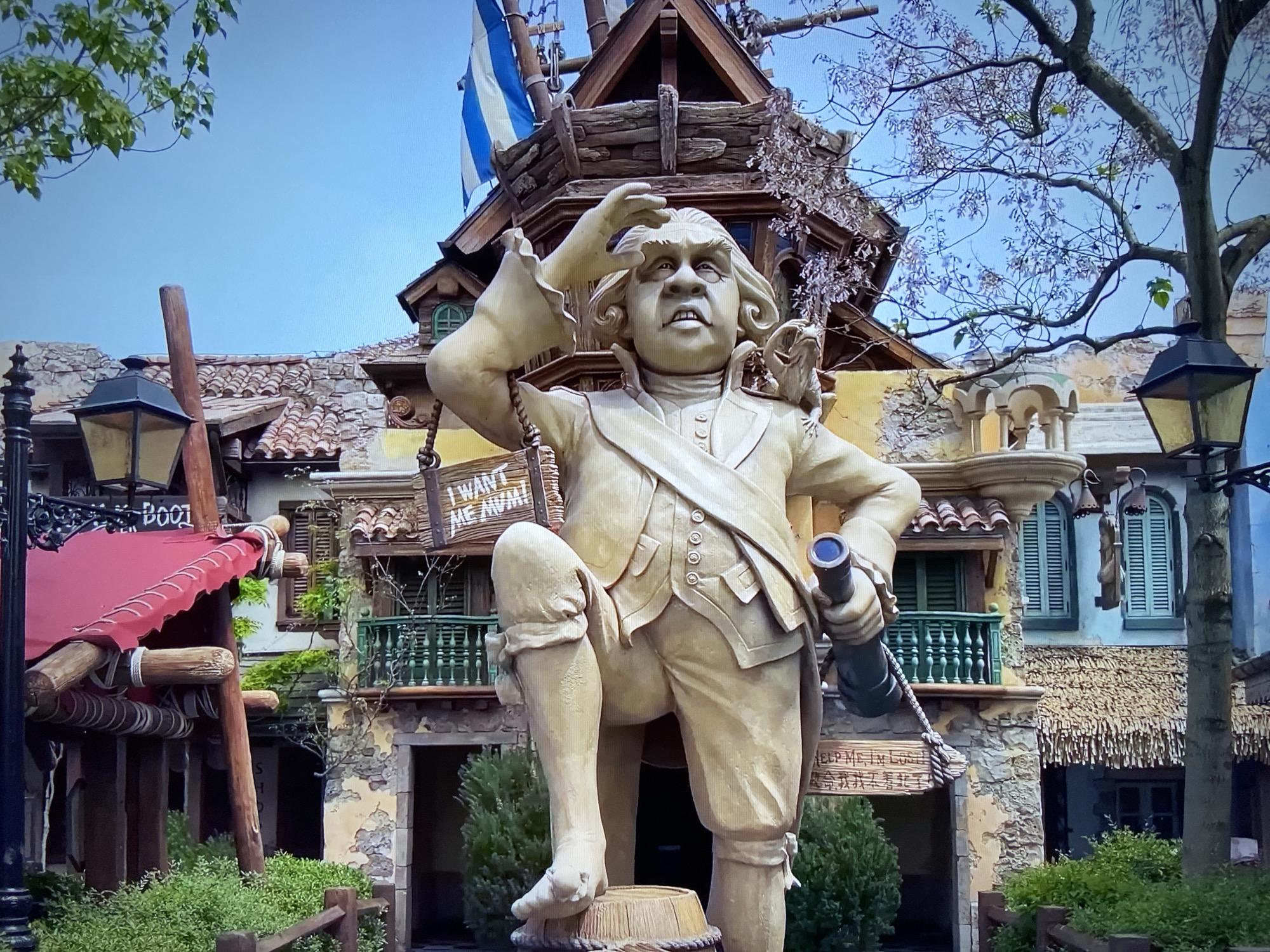 Captured from a low angle, the photograph showcases a sculpted figure positioned prominently in front of a themed building. The sculpted figure, likely depicting a historical figure, wears period clothing, including a jacket, breeches, and buckled shoes. The figure is rendered in a light, possibly stone-like material.
Behind the figure, a building with a fantastical design rises. The architecture incorporates elements of wood and stone, with multiple levels and decorative details. A flag with alternating blue and white stripes adorns the upper part of the structure.
To the sides, trees with emerging foliage frame the scene, hinting at spring or early summer. Black lampposts stand on either side of the frame, adding to the scene's ambiance. A sign with lettering is visible on the left side of the structure.
The photograph's composition and elements suggest the scene is located within a themed park or attraction.