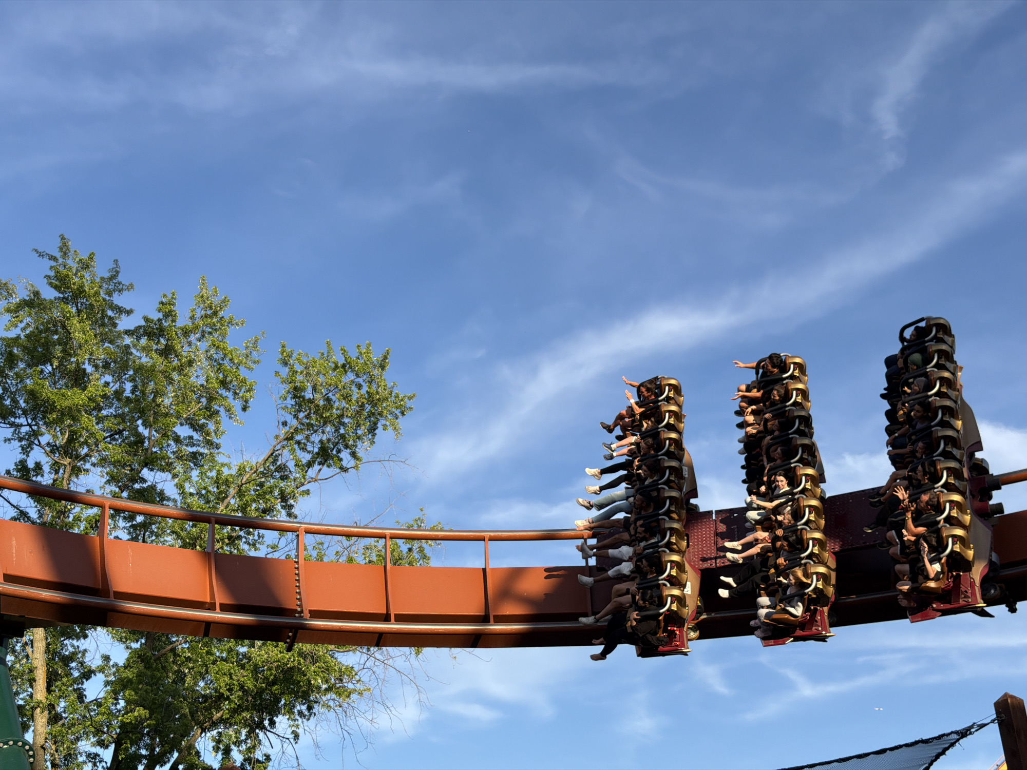 A roller coaster car carrying numerous passengers is in motion against a clear blue sky with scattered wispy clouds. The roller coaster track, painted a bright orange, curves to the left in the foreground, bordered by green trees. The passengers are seated in the car, some with arms raised in the air, indicating excitement or exhilaration. The roller coaster appears to be suspended or at a high point in its track. This scene is located at Canada's Wonderland in Vaughan, Ontario, Canada, an amusement park known for its thrill rides.