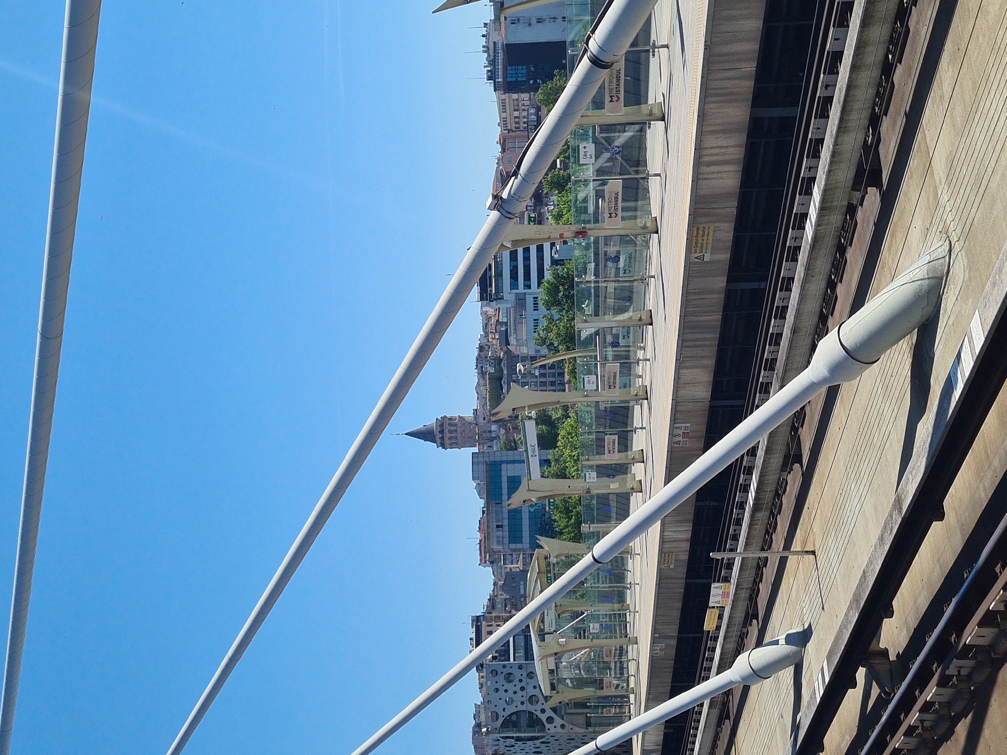 Here's a description of the image:
The image appears to be taken from a structure, likely a bridge, looking out over a cityscape. The foreground features prominent white structural elements, possibly suspension cables or support beams of the bridge itself. The background reveals a dense urban setting with various buildings, including what seems to be a historical tower or spire dominating the skyline. There are also modern buildings with glass facades interspersed among the older structures.
The presence of a Metro Istanbul platform and signs suggests that this is most likely located in Istanbul, Turkey.