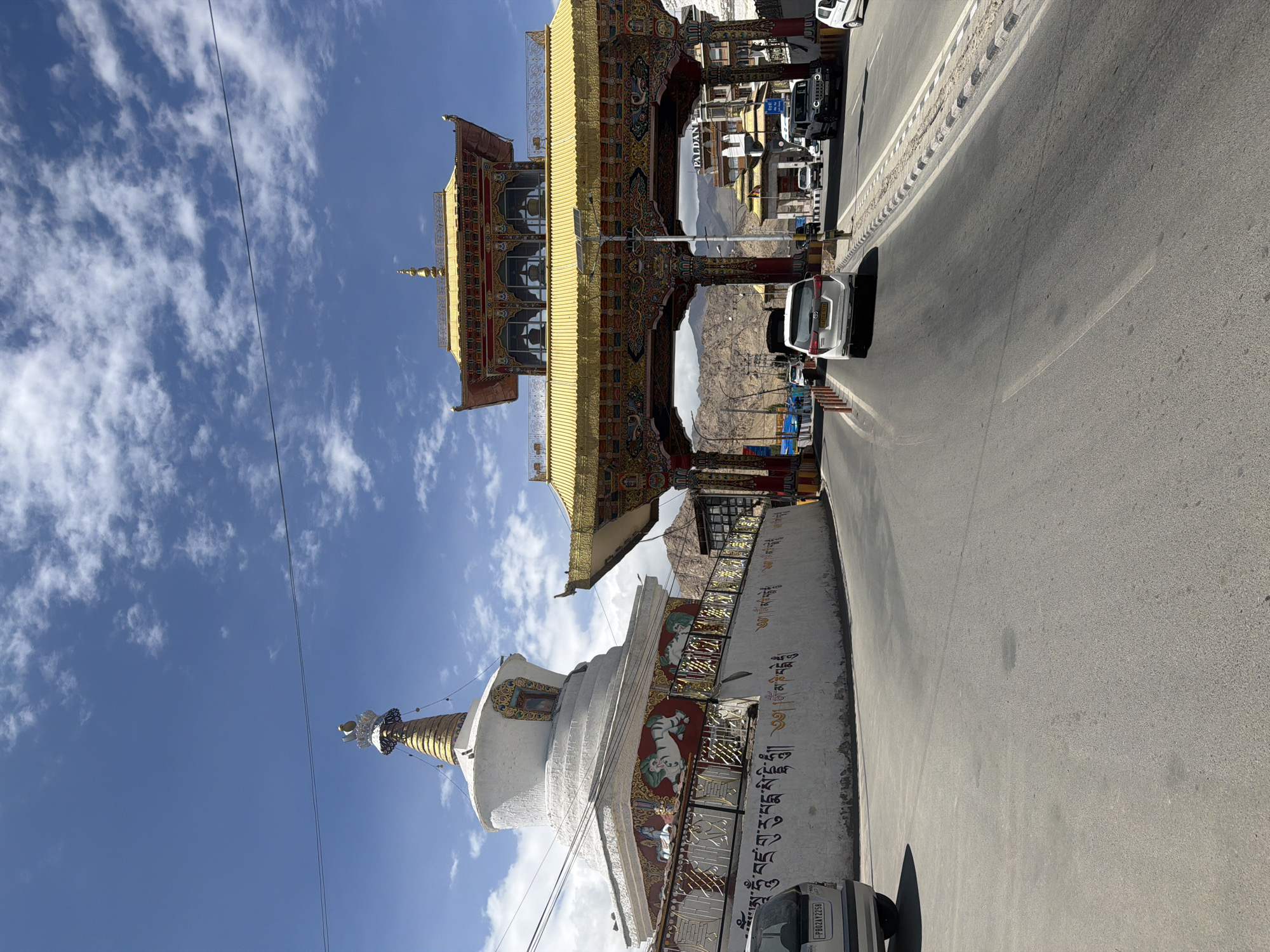 This image captures a street scene in Leh, India, featuring a prominent stupa on the left and a vibrantly decorated golden archway on the right. The stupa, a white dome-shaped structure with a golden spire, is adorned with intricate murals and Tibetan script along its base. The archway, characterized by its golden roofs and multi-colored ornate carvings, serves as an entrance to a complex that includes buildings with the signage "Paldan". Several vehicles, including cars and a jeep, are visible on the paved road, which curves through the scene. The environment is arid, with mountains visible in the background under a bright blue sky with scattered clouds. The overall impression is one of a culturally significant and bustling urban area in a mountainous region.
