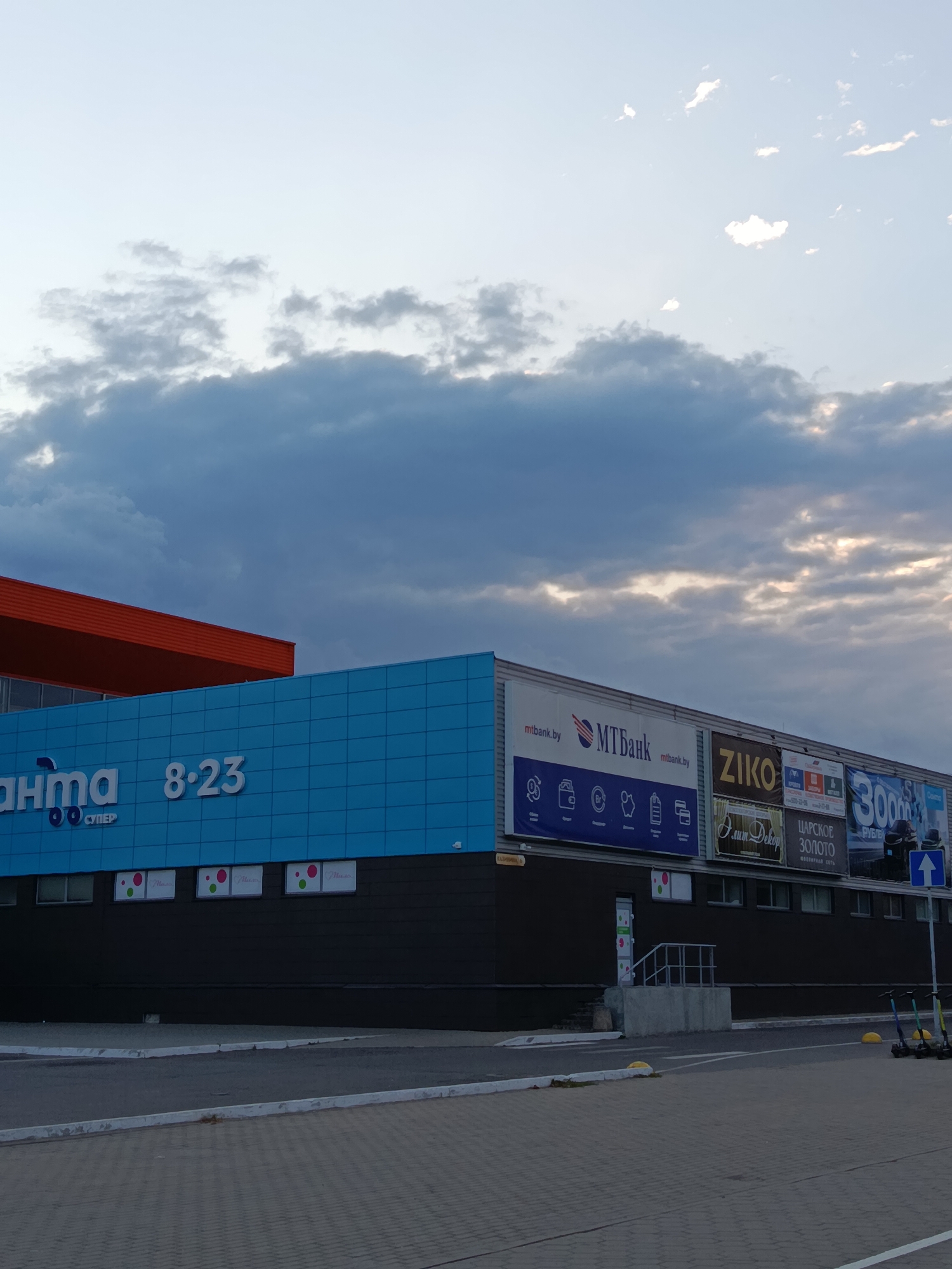 The image shows the exterior of a commercial building with a distinctive bright blue tiled facade. Above the blue tiles, an orange awning is visible. The building has several large advertisements mounted on its upper section. One advertisement prominently displays "MTBank.by" and the MTBank logo, along with icons representing financial services. Another advertisement features "ZIKO". Further to the right, there are advertisements for "V.sum Dekor", "ЦАРСКОЕ ЗОЛОТО" (Tsarskoye Zoloto), and another large banner with the text "30000". A few people are visible near the right edge of the building, one of whom is wearing a bright blue shirt and standing near a white directional arrow sign. In the foreground, there is a paved area with yellow bollards and several electric scooters parked to the right. The sky above is filled with dramatic, dark clouds, with patches of sunlight breaking through. The overall scene suggests a commercial area in Svietlahorsk, Belarus, likely in the late afternoon or early evening given the lighting.