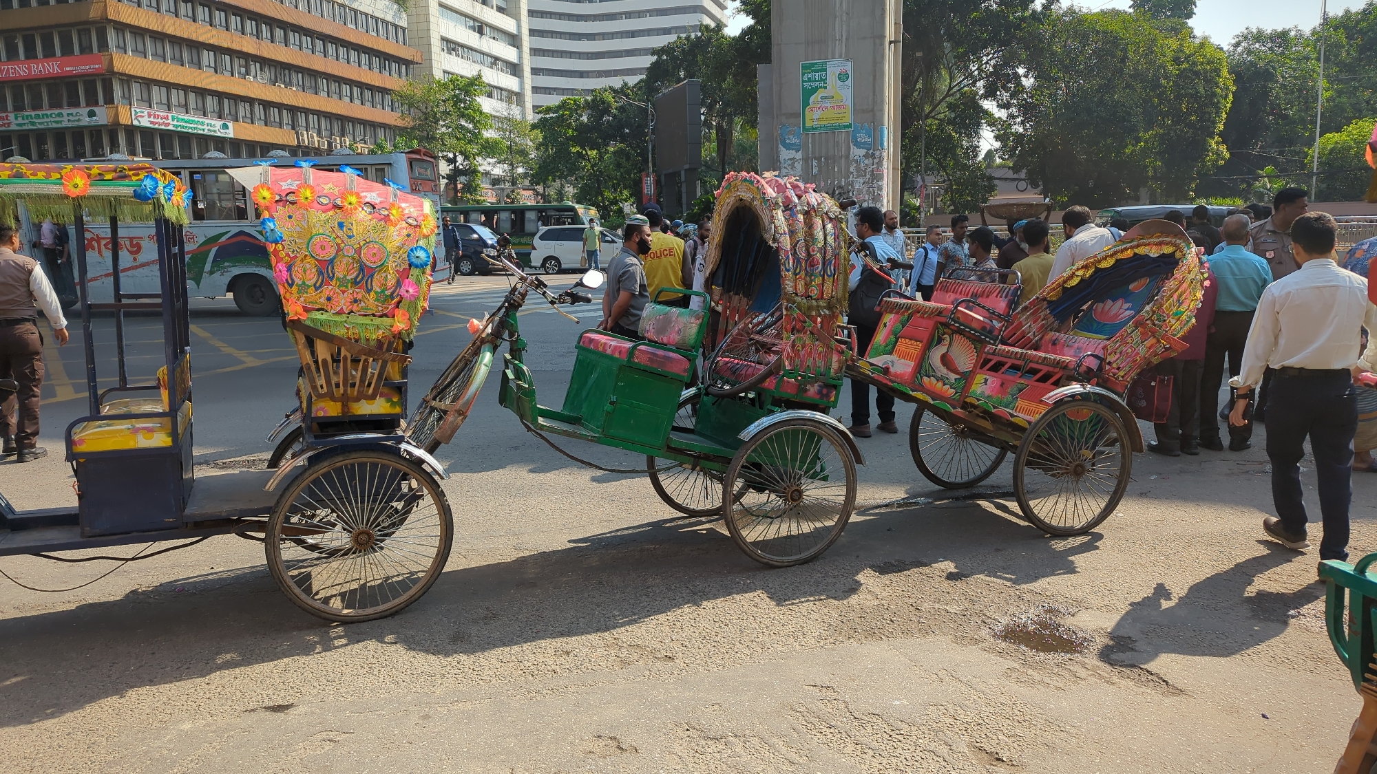 The image captures a typical urban street scene in Dhaka, Bangladesh, featuring a line of parked rickshaws. These cycle rickshaws, a common mode of transportation, are brightly decorated with colorful artwork and ornamentation. A fourth rickshaw is hitched behind the first. Several people, including a police officer in uniform and civilians, are standing nearby and appear to be in conversation. The scene is set against a backdrop of tall buildings, suggesting a busy commercial area. Street signs and posters on a pillar are also visible in the distance. The lighting suggests it is daytime. The overall impression is that of a crowded and bustling urban environment in Dhaka.