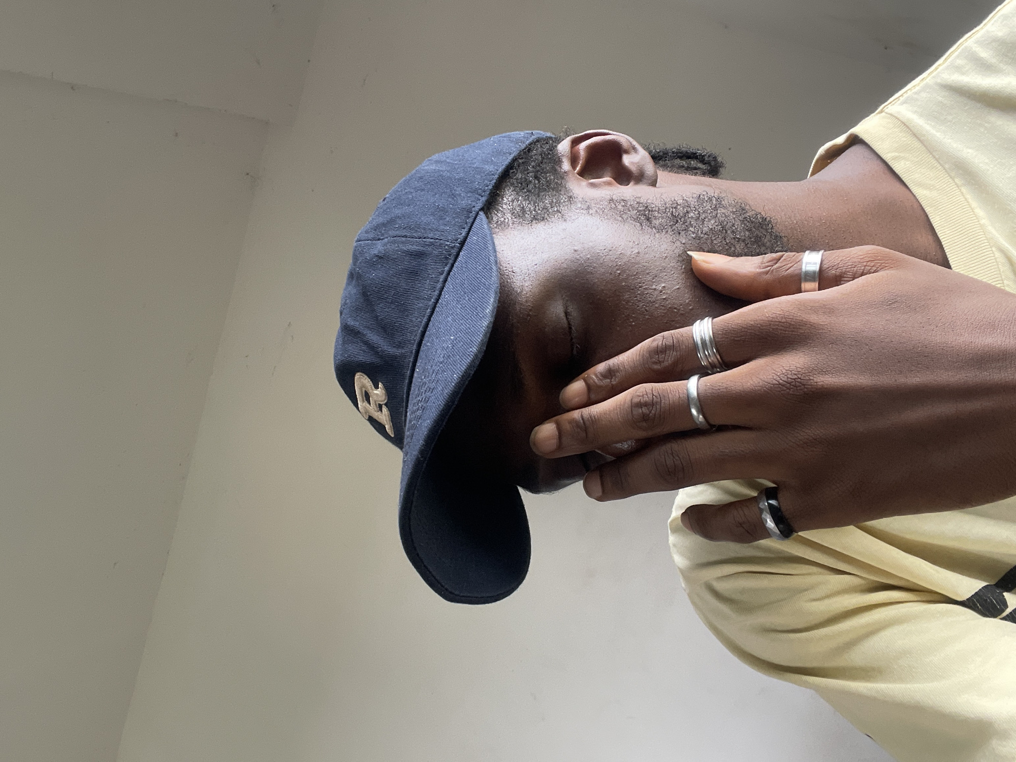 The image shows a close-up, head-and-shoulders view of a person wearing a navy blue baseball cap with a gold embroidered "R" and a light yellow t-shirt. The person's right hand is covering their face, obscuring their eyes. They are wearing several silver rings on their fingers, including a faceted black and silver band on their left ring finger, and plain silver bands on their middle and ring fingers of their right hand. The background appears to be an indoor setting with neutral-colored walls. There are no overt indications of the specific location within Abuja, Nigeria, beyond the implied domestic or private setting suggested by the background.