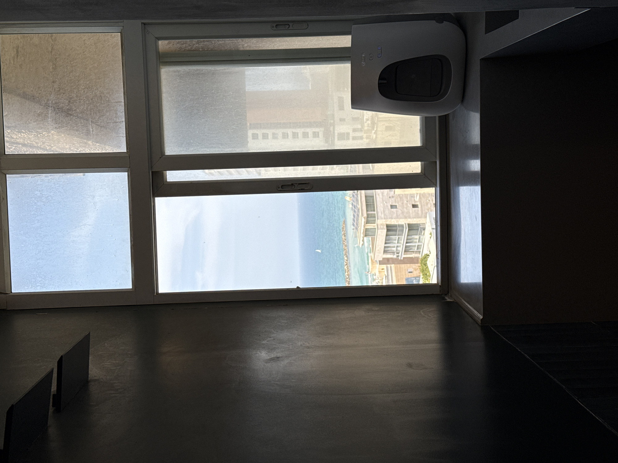 A low-angle shot shows an interior space with a large window overlooking the city of Tel Aviv, Israel and the Mediterranean Sea. The window is comprised of several panes, some with significant condensation or dirt obscuring the view. Through the clear panes, one can see the coastline, the sea, and buildings characteristic of the Tel Aviv cityscape. A robotic window cleaner is attached to the upper right section of the window. The interior floor is a dark, reflective surface. A section of a dark-colored couch or seating is visible in the lower-left corner. The right side of the frame is shrouded in shadow, revealing a wall and floor.