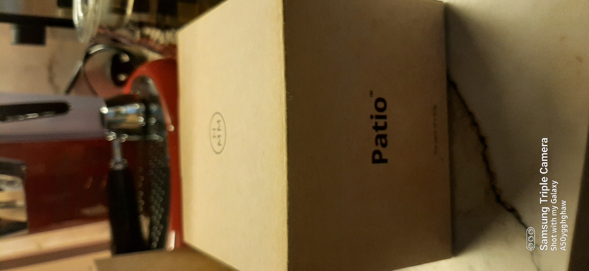 The image shows a close-up of a cardboard box with the word "Patio" printed on it. Above the word "Patio," there is a small oval logo with the letters "H" and "MM" inside. The box appears to be placed on a marble surface. In the background, out of focus, are parts of what look like kitchen appliances, possibly a red espresso machine and a silver kettle. The lighting is dim, casting shadows. The bottom of the image shows a watermark from a Samsung camera, indicating the photo was taken with a Samsung Galaxy A50. There are no people or animals visible in the frame. The location is not specified in the image or its metadata.