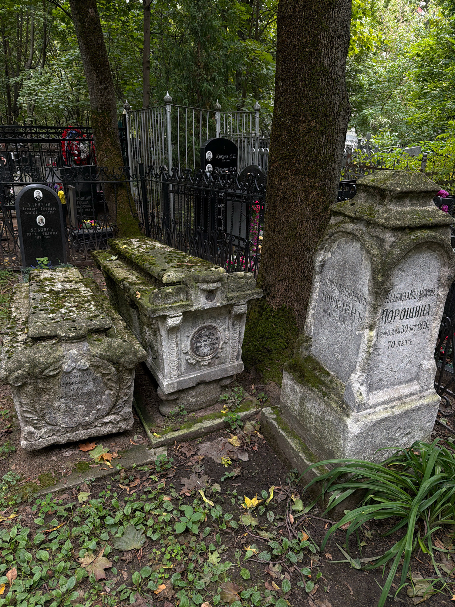 The image depicts a section of the Novodevichy Cemetery in Moscow, Russia. The scene features several tombstones and crypts, some of which are heavily weathered and covered in moss. In the foreground, two stone crypts, resembling sarcophagi, are partially visible, with ornate carvings and moss growth. To the right, a tall, upright tombstone with inscriptions in Cyrillic is prominent. The inscriptions on this tombstone include names and dates, one reading "АЛЕКСАНДРА МИХАЙЛОВНА ПОРОШИНА" (Aleksandra Mikhailovna Poroshina) and mentioning her death on "30 ОКТЯБРЯ 1871" (October 30, 1871) at "70 ЛЕТЪ" (70 years). Another inscription to the left appears to be for "ПОРФИРИЙ ТЕРЕНТЬЕВИЧ" (Porfiry Terentyevich). Behind the tombstones, a dark, wrought-iron fence encloses the cemetery plots, with trees and dense foliage visible in the background, suggesting an established, overgrown cemetery setting. The ground is covered with fallen leaves, soil, and patches of green ground cover and grass. The overall atmosphere is one of age, history, and solemnity, characteristic of an old cemetery.