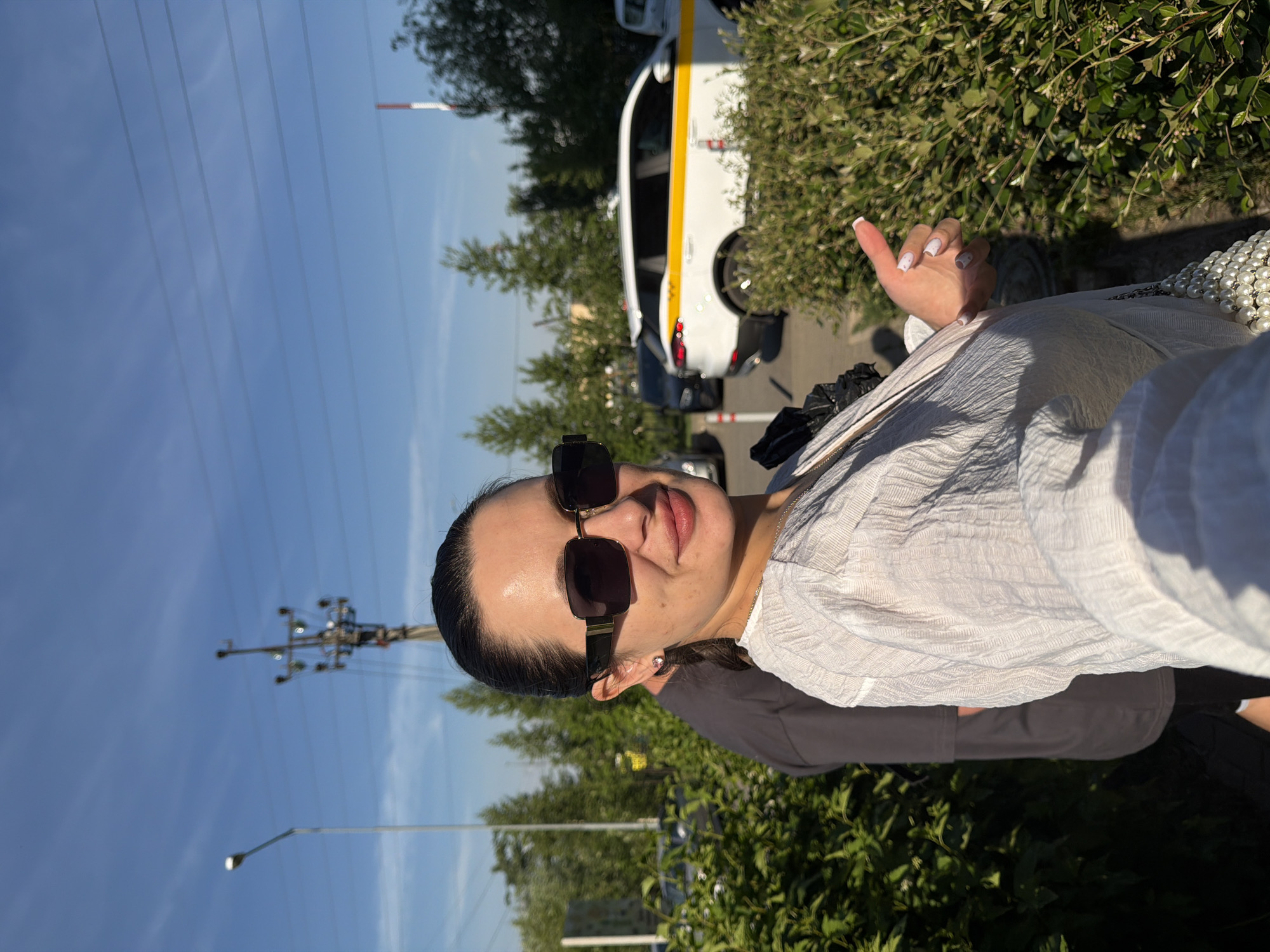 The image depicts an outdoor scene in an urban or suburban environment, consistent with the city of Odintsovo, Russia, given the identifiable Yandex Taxi. A woman is positioned slightly left of center, visible from the chest up, looking directly at the camera with a slight smile. She has dark hair pulled back, wears dark rectangular sunglasses with gold-colored accents on the temples, and a light-colored, textured, possibly crinkled, long-sleeved top. A small stud earring is visible in her left ear. Her right hand is present in the lower right, displaying white-tipped fingernail polish with small pink dots, and her thumb is pointing upwards. A pearl-beaded accessory is visible hanging from her shoulder in the lower right. A second individual is partially obscured behind and to the left of the woman, wearing a dark grey top.
The background features a clear blue sky traversed by multiple parallel power lines. A tall street lamp stands on the far left. A complex utility pole with numerous crossarms and insulators is prominently located slightly left of the center. On the right side of the mid-ground, a white SUV with a distinct yellow stripe, identifiable as a Yandex Taxi (likely a Geely Atlas Pro or similar model), is parked. Other vehicles, including a dark-colored car behind the Yandex Taxi and a silver car further left, are partially visible through the foliage. Lush green bushes occupy the foreground and mid-ground. A red and white striped pole is situated on the right side of the paved area, behind the bushes. Various trees are scattered throughout the background. The scene is brightly illuminated by natural sunlight, casting shadows.