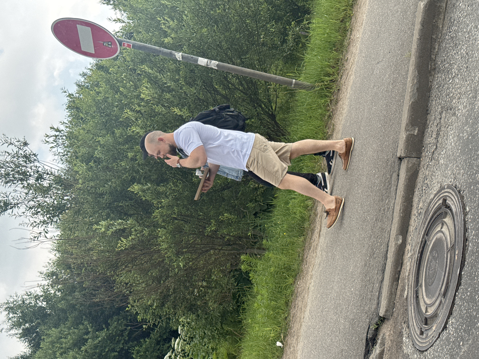 This media file shows two people outdoors near a road and a grassy area with trees. The person in the foreground is a man with a shaved head, wearing a white t-shirt and beige shorts. He is holding a phone to his face and appears to be looking at it. He is also wearing brown boat shoes. Behind him, another person is visible wearing a black backpack and part of a blue denim jacket. They are also holding a flat, light-colored object, possibly a tablet or a book.
The scene appears to be a sidewalk or path next to a paved road. A concrete curb separates the road from the grassy verge. In the background, there are dense green trees and foliage. A metal pole with a red and white circular sign is visible above and to the left of the man. The sky is overcast with clouds. The lighting suggests it is daytime. The overall impression is casual, possibly a break or a moment of rest during an outing.
There is no visible text in the media file other than on the road sign, which is a standard European "no entry" sign indicating a prohibition of entry for all vehicles, with a white rectangle in the center. The context provided places the location in Novoe Devyatkino, Russia.