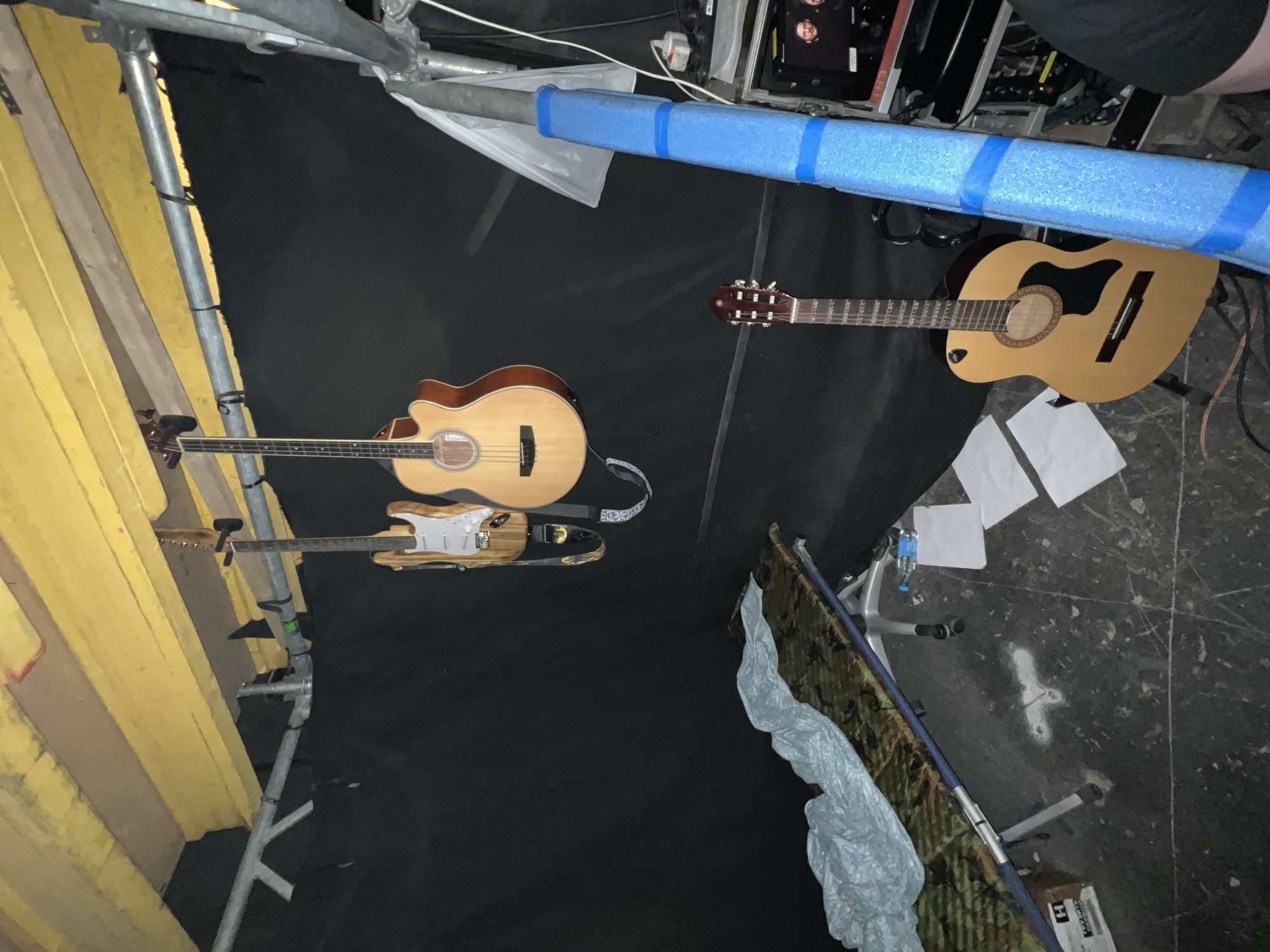 This image captures a utilitarian backstage or preparation area for a musical event in Khimki, Russia. The scene features three string instruments laid on a dark, textured floor. An acoustic bass guitar with a light wood finish and a cutaway body is positioned on the left, next to an electric guitar with a pale body and a wooden fretboard. To the right, a standard acoustic guitar with a natural finish and classical tuning pegs rests near the top of the frame.
Prominently featured are various elements of temporary construction or stage setup. On the left, a tall yellow scaffolding structure and wooden planks frame the edge of the scene. Across the top, a horizontal metal pole, likely part of the scaffolding, is wrapped in bright blue foam padding secured with blue tape. Below this, wires and cables are visible, typical of audio or lighting equipment.
A person's leg and lower torso, wearing dark clothing, are partially visible at the top right, indicating human presence. Several pieces of white paper are scattered on the floor near the acoustic guitar. In the bottom right corner, a grey tarp or sheet covers an unseen object, adjacent to a folded camouflage-patterned fabric item. A clear plastic water bottle stands near a metal stand or chair base. A cardboard box in this area has the letter "H" visible on its side.
The lighting is dim, suggesting an indoor setting or nighttime. The overall impression is of a functional space for musicians, either during setup, a break, or after a performance.