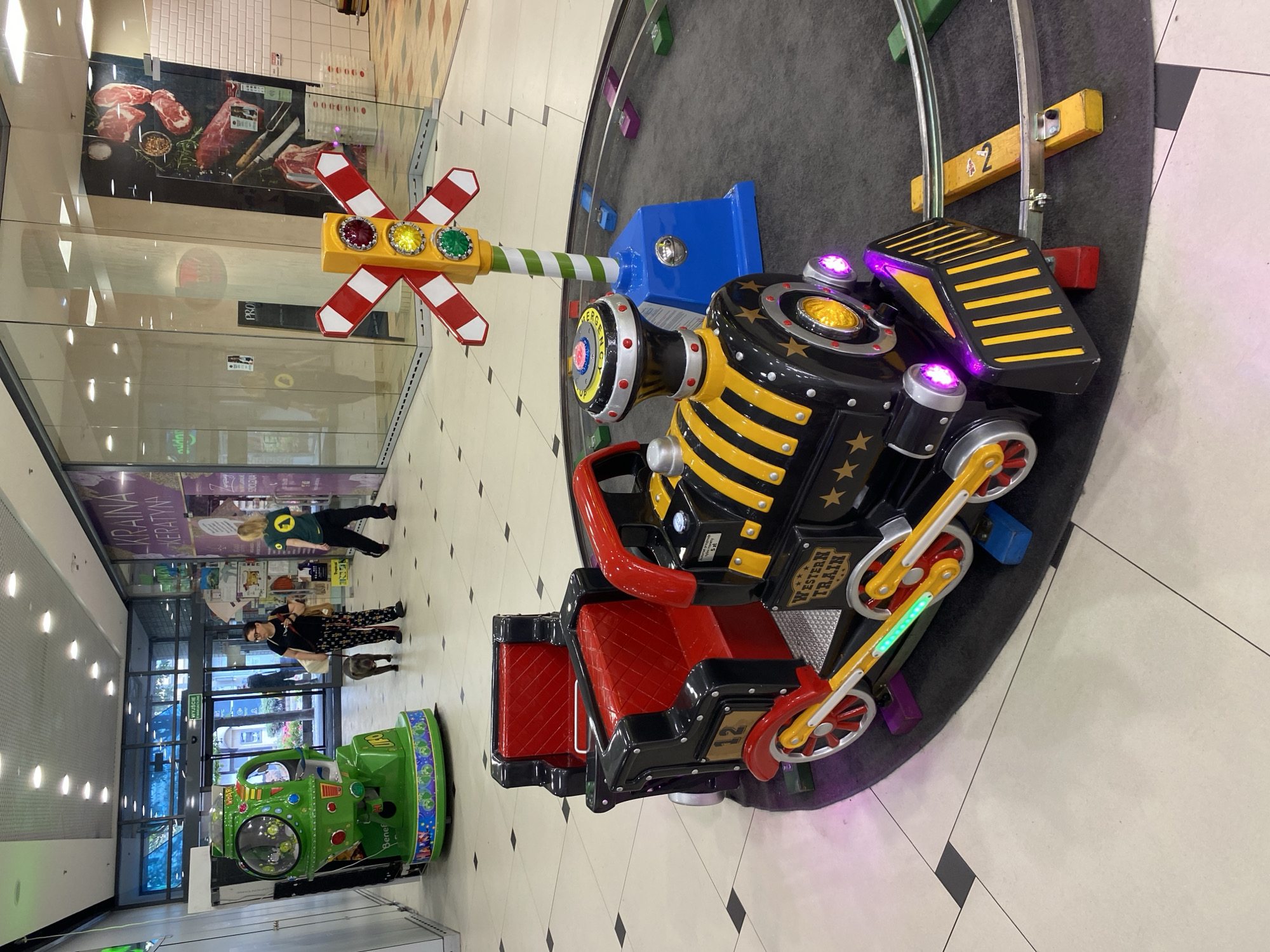 The image depicts a scene inside a shopping mall, likely in Szczecin, Poland, with a focus on a children's train ride. In the foreground, a black and yellow toy train with red seating is positioned on a circular track. The train has "WESTERN TRAIN" written on its side and the number "12" on its wheel. To the right of the train is a colorful traffic signal decoration with red, yellow, and green lights, and a railroad crossing sign. The track is made of metal and supported by colorful blocks. In the background, a green UFO-themed ride is visible to the left, with a sign that partially reads "Benef..." and "Dorell". Further in the background, two women and a dog are walking. One woman, wearing a black top and patterned pants, is walking a gray dog on a leash. The other woman is wearing a green t-shirt with a circular logo and dark pants. They are walking past shops with glass fronts, one of which displays an advertisement for "KRAINA KERATYNA". The mall floor is tiled in a checkered pattern of beige and dark gray squares. The ceiling features recessed lighting.