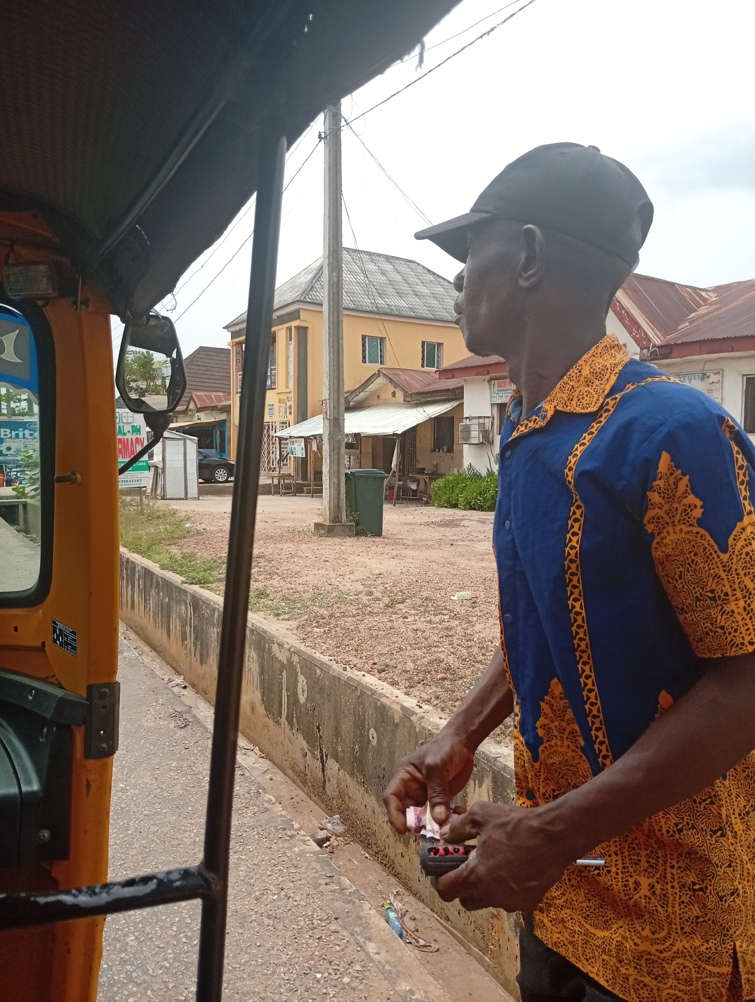 A man of dark complexion is depicted standing outdoors, in profile, next to an orange and blue auto-rickshaw (keke napep) in Adankolo, Nigeria. He wears a black baseball cap and a short-sleeved blue shirt adorned with intricate yellow-orange patterned designs on the collar, shoulders, and down the front. His hands are positioned in front of him, holding what appears to be Nigerian Naira notes alongside a small, dark, possibly plastic, multi-compartment item.
The setting is a roadside under an overcast daytime sky. To the left, part of the auto-rickshaw is visible, including a side mirror. A blue sign on the auto-rickshaw displays "Brit" and a partial phone number "08053652182". The ground is covered with dirt and gravel, bordered by a concrete wall or gutter. In the background, various buildings with tin roofs are scattered, including a prominent yellow-orange two-story structure. A white sign on a building in the mid-ground reads "AL-PHARMACY". A green refuse bin and a utility pole are also visible. The man's expression is neutral, focused on the task at hand.