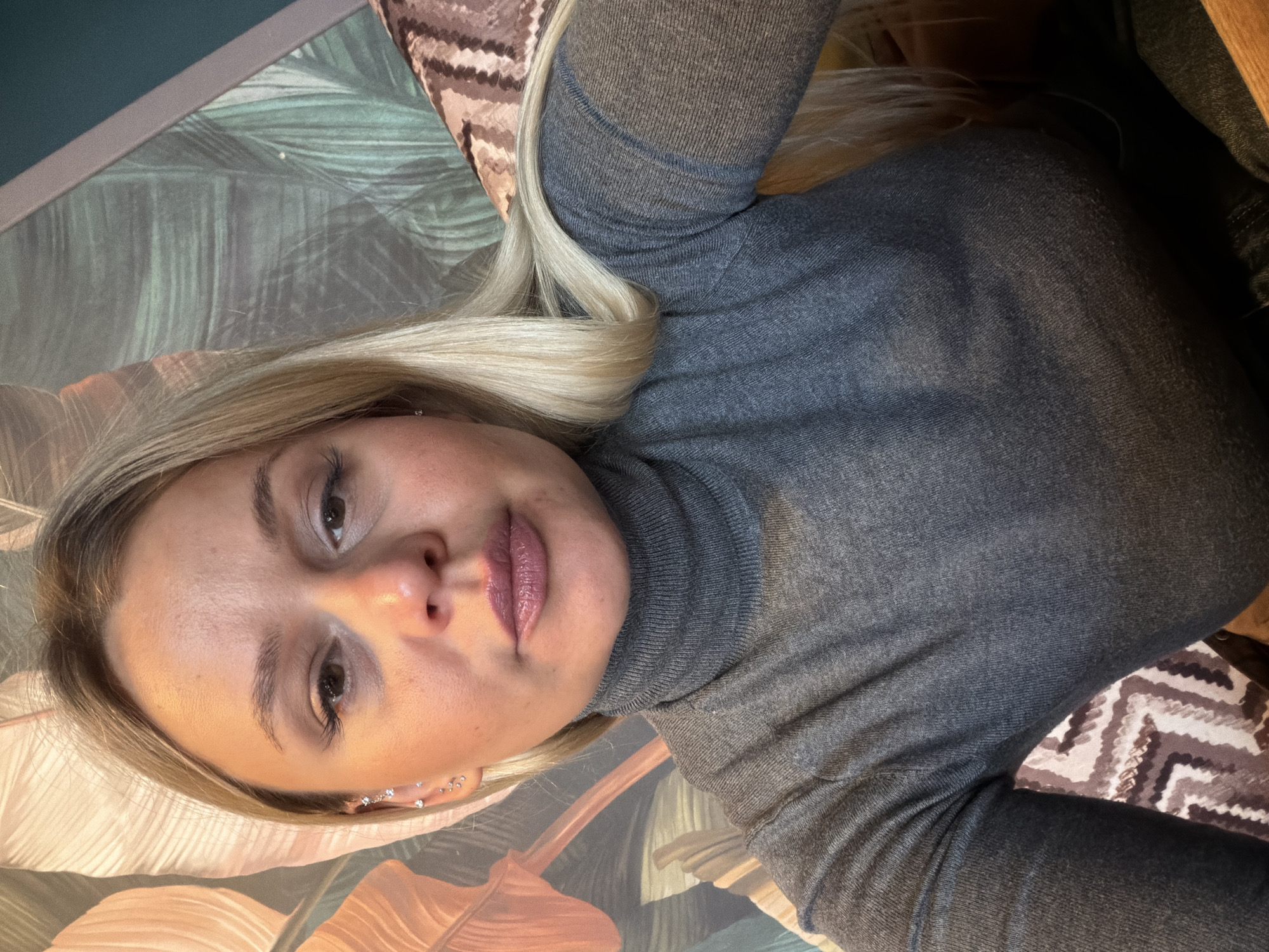 The image shows a blonde woman with a grey turtleneck sweater. She is close to the camera and is facing the viewer. The background includes a green and brown leafy design and a brown and white zigzag pattern. The woman appears to be wearing multiple earrings. The location of Sarapul, Russia is not discernable in the image.